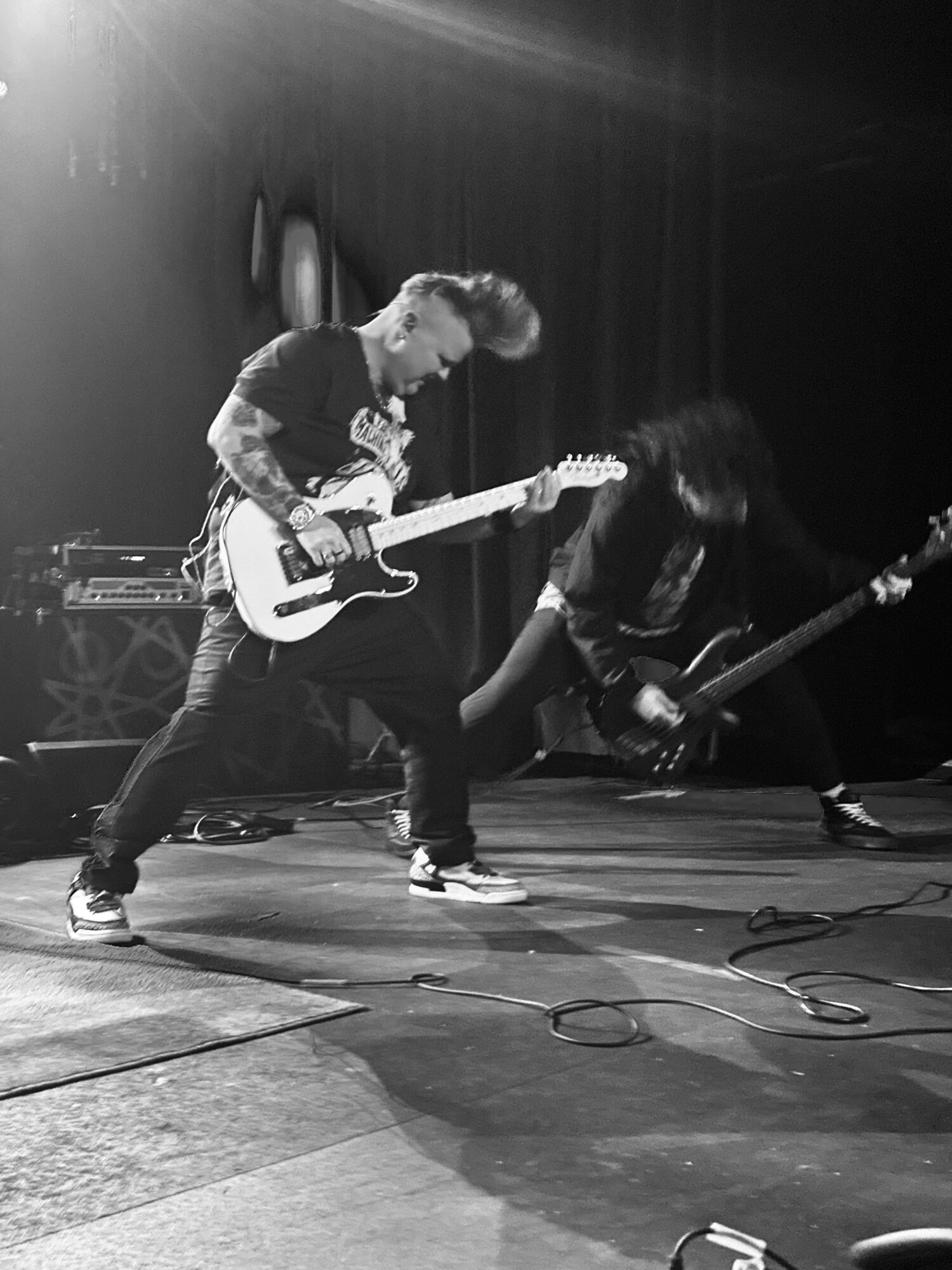 Two musicians playing electric guitars on stage, one standing and the other crouching, with stage equipment visible.