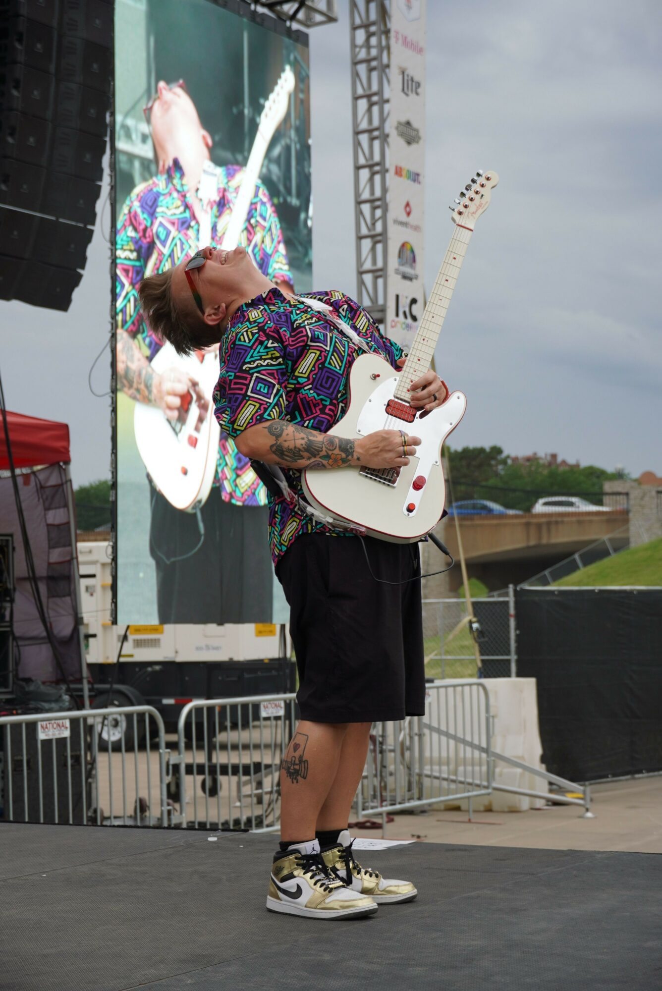 Person playing guitar on stage with a large screen behind, wearing colorful clothing and sunglasses.