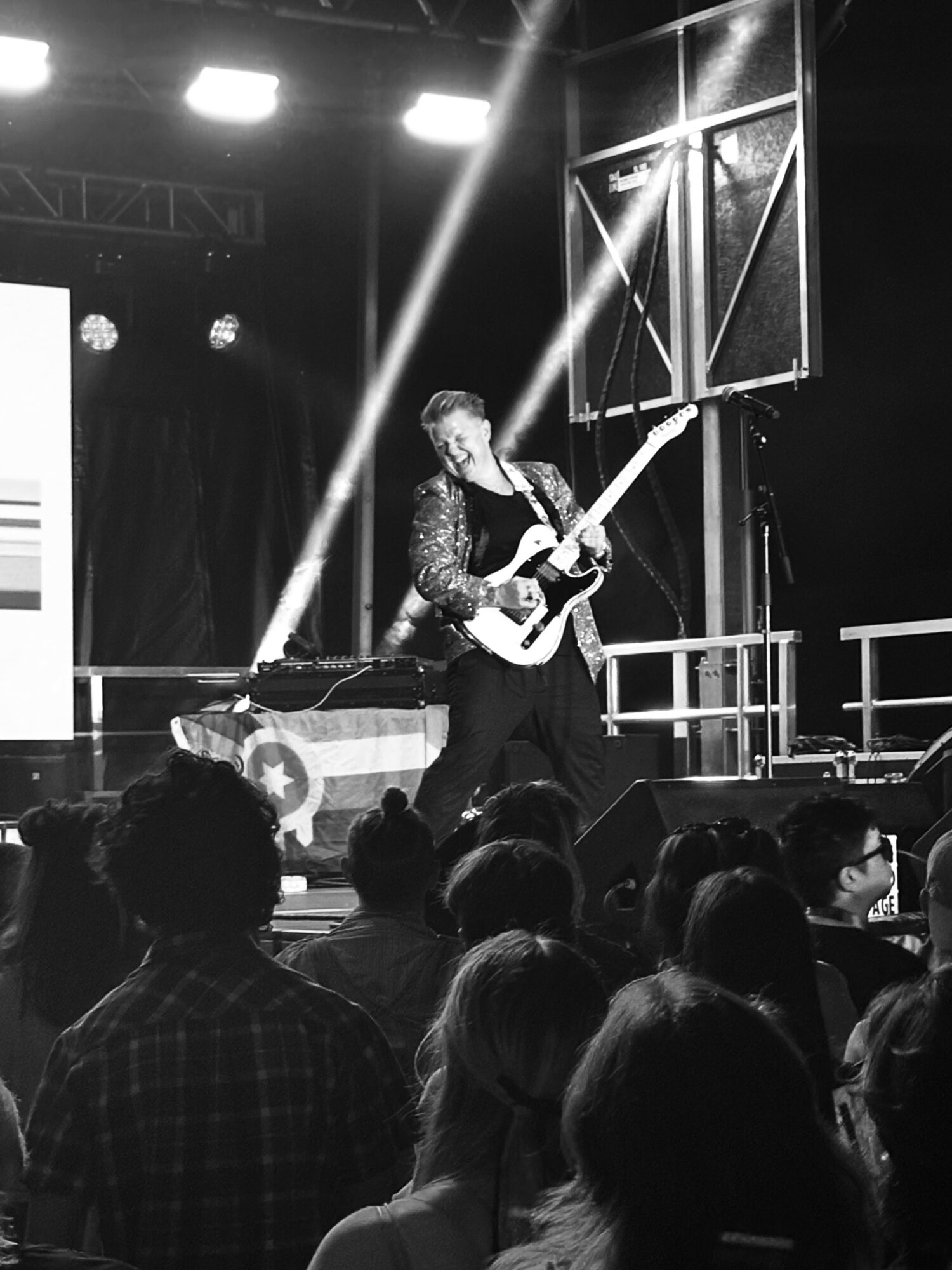 Musician playing guitar on stage with audience in foreground, stage lights and equipment visible, black and white image.