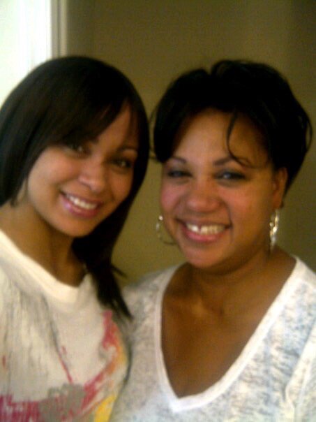 Two women smiling, one with shoulder-length dark hair and the other with short dark hair and hoop earrings, indoors.