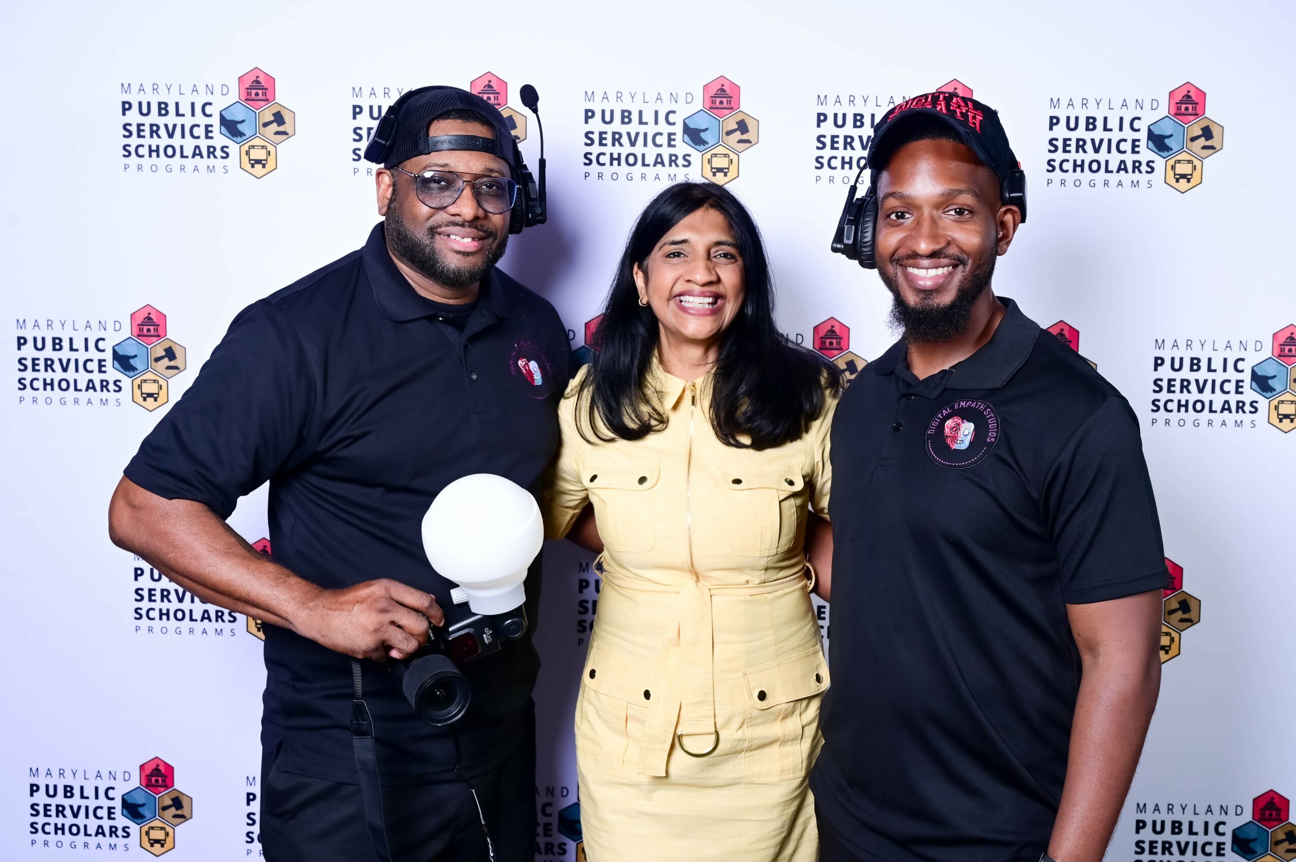 Three people smiling, standing in front of a backdrop with logos, two men and one woman, all dressed casually.
