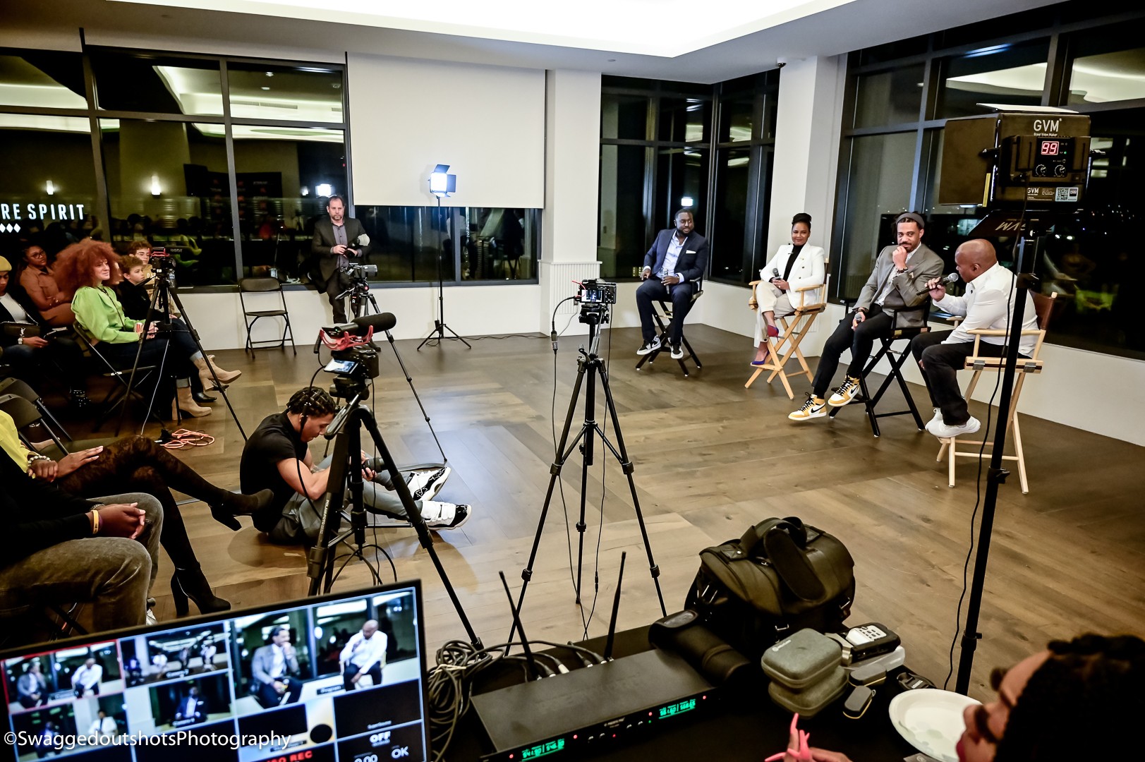 Panel discussion with five people seated on stage, audience watching, cameras and equipment in foreground, large windows behind.