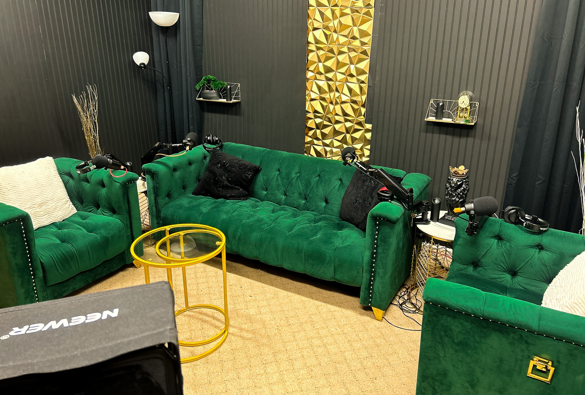 Three green velvet sofas in a room with black walls and gold decorative panel, with yellow side table and various objects around.