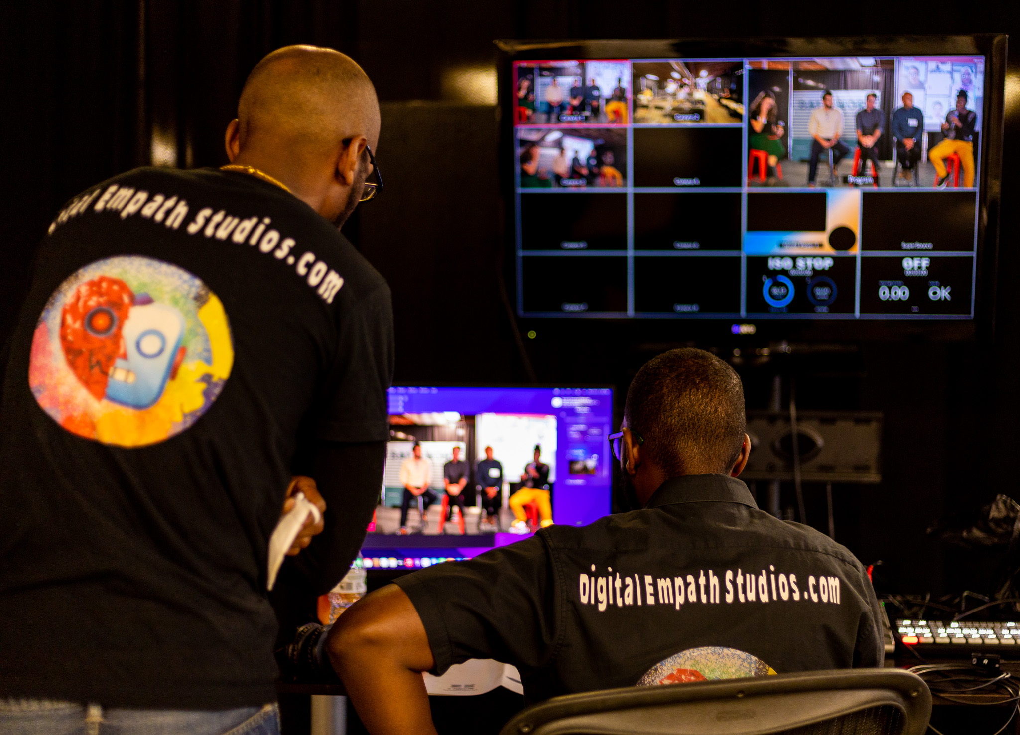 Two people work in a control room with multiple monitors displaying video feeds, one standing and one sitting, both wearing black shirts.
