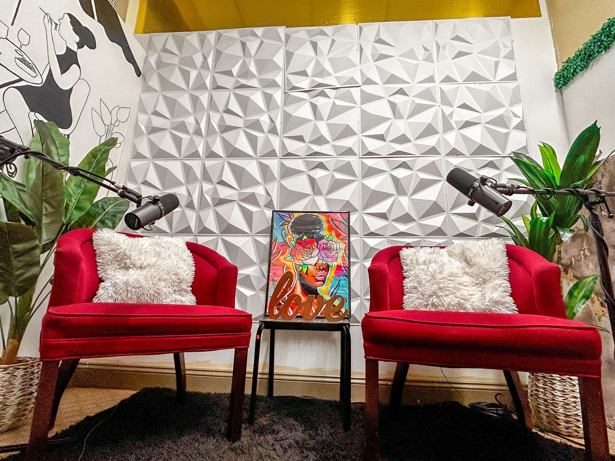 Two red velvet sofas with white pillows face each other, with a colorful artwork between them, against a textured white wall.