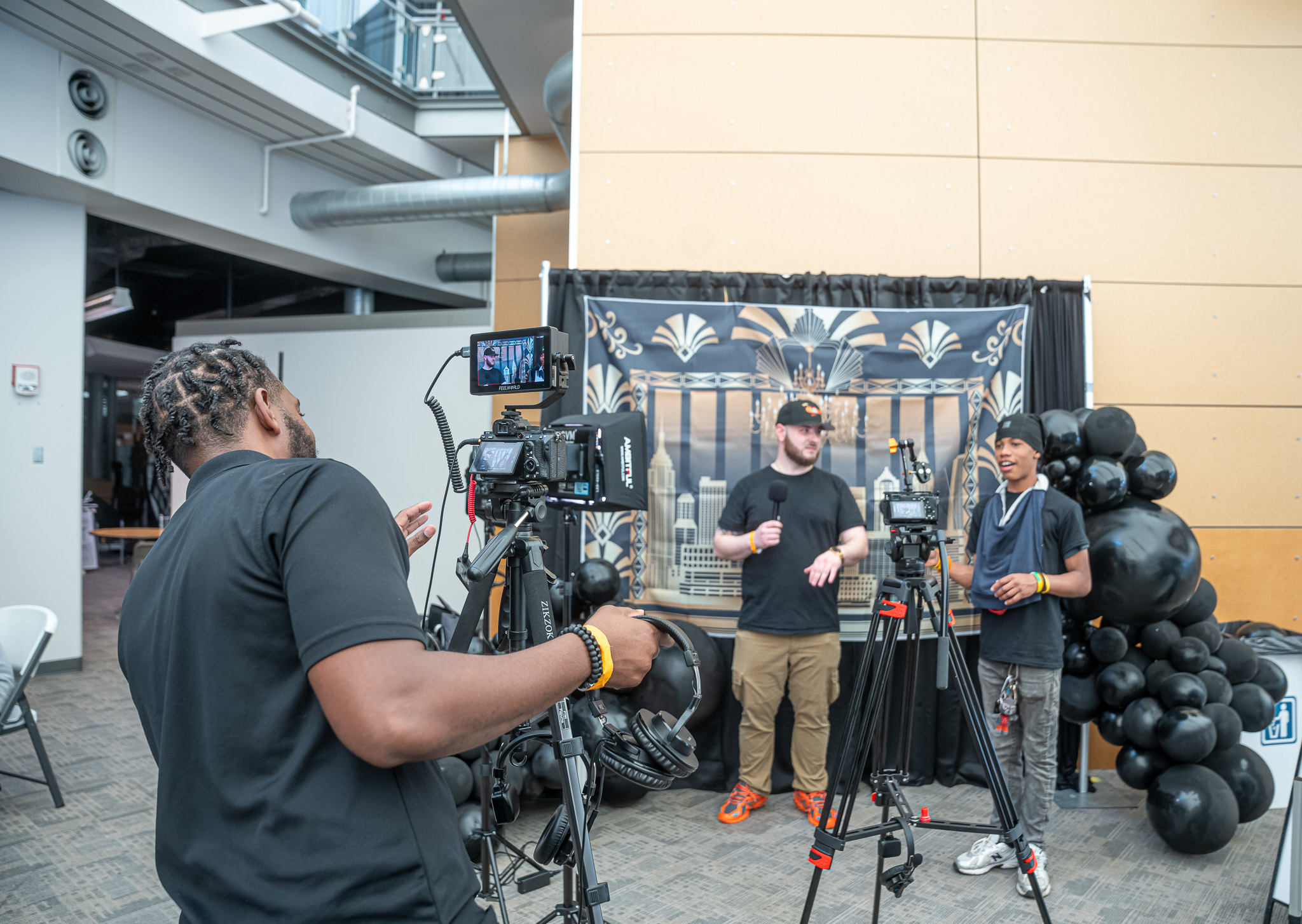 Two men are being filmed by a cameraman in front of a decorated backdrop with black balloons, in an indoor setting.