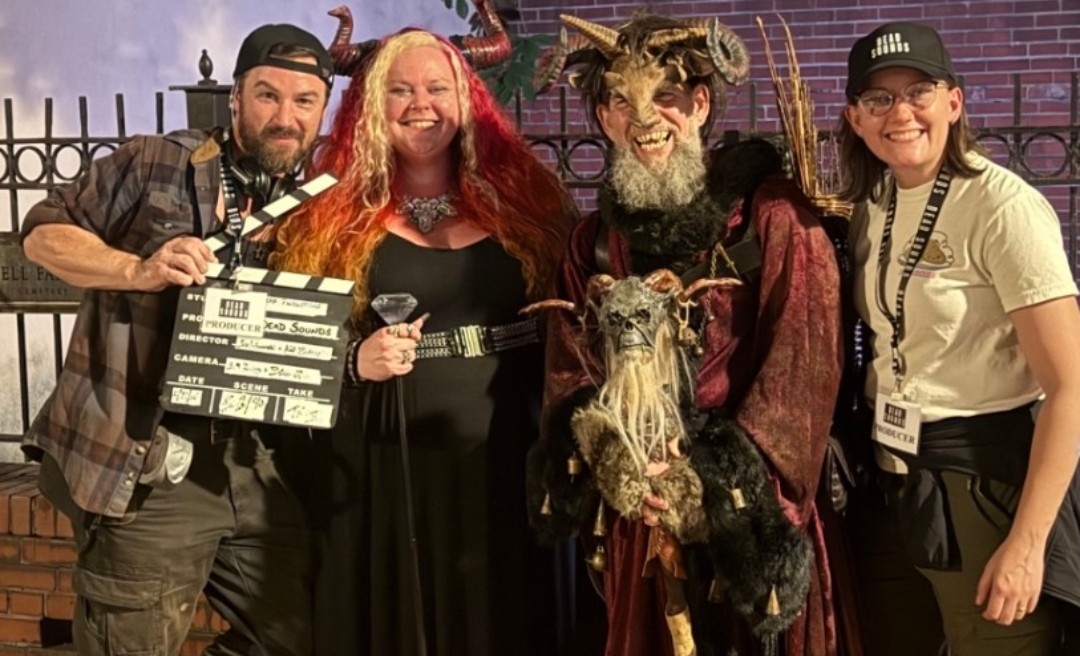 Four people in costumes and casual clothing pose outdoors at night, with a fence and city lights in background.