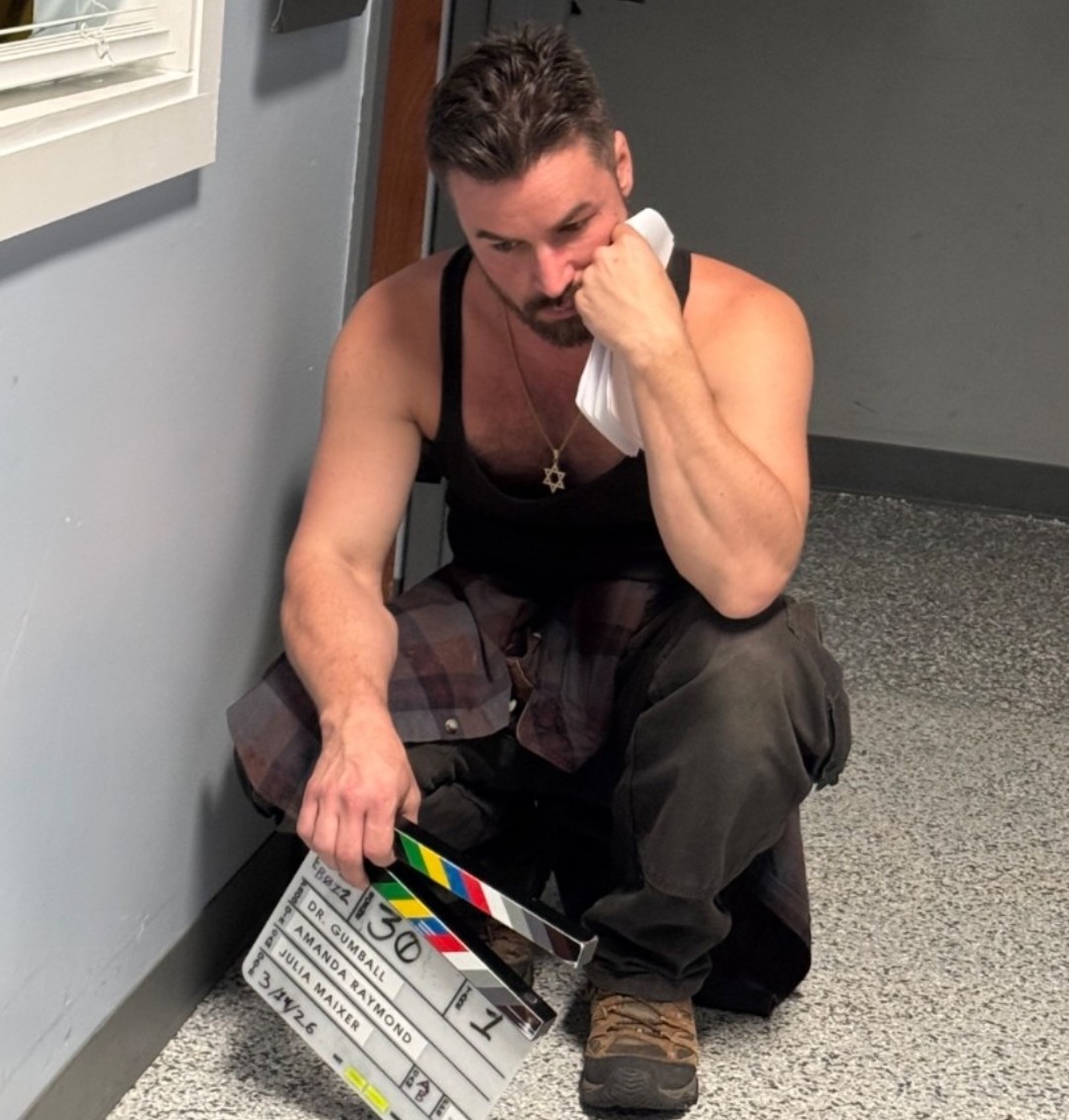 Man sitting on the floor, resting his head on his hand, holding a clapperboard, in a room with gray walls and carpet.