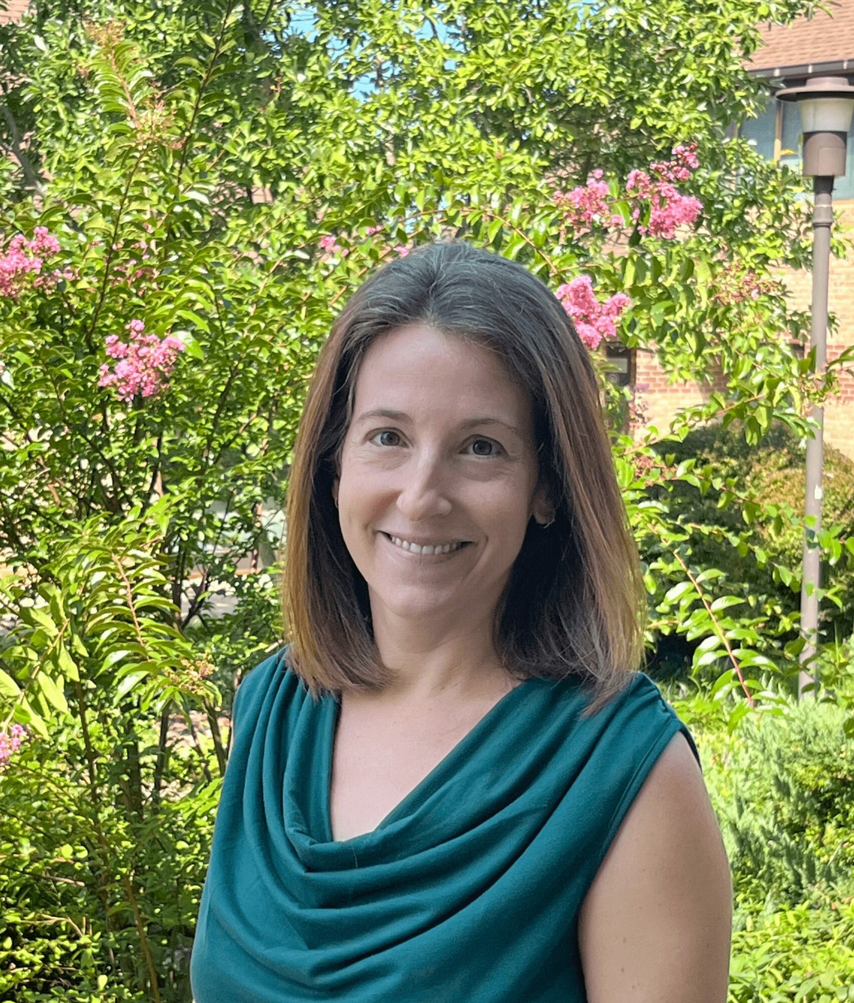 Woman with shoulder-length hair smiling outdoors with green trees and pink flowers in background.