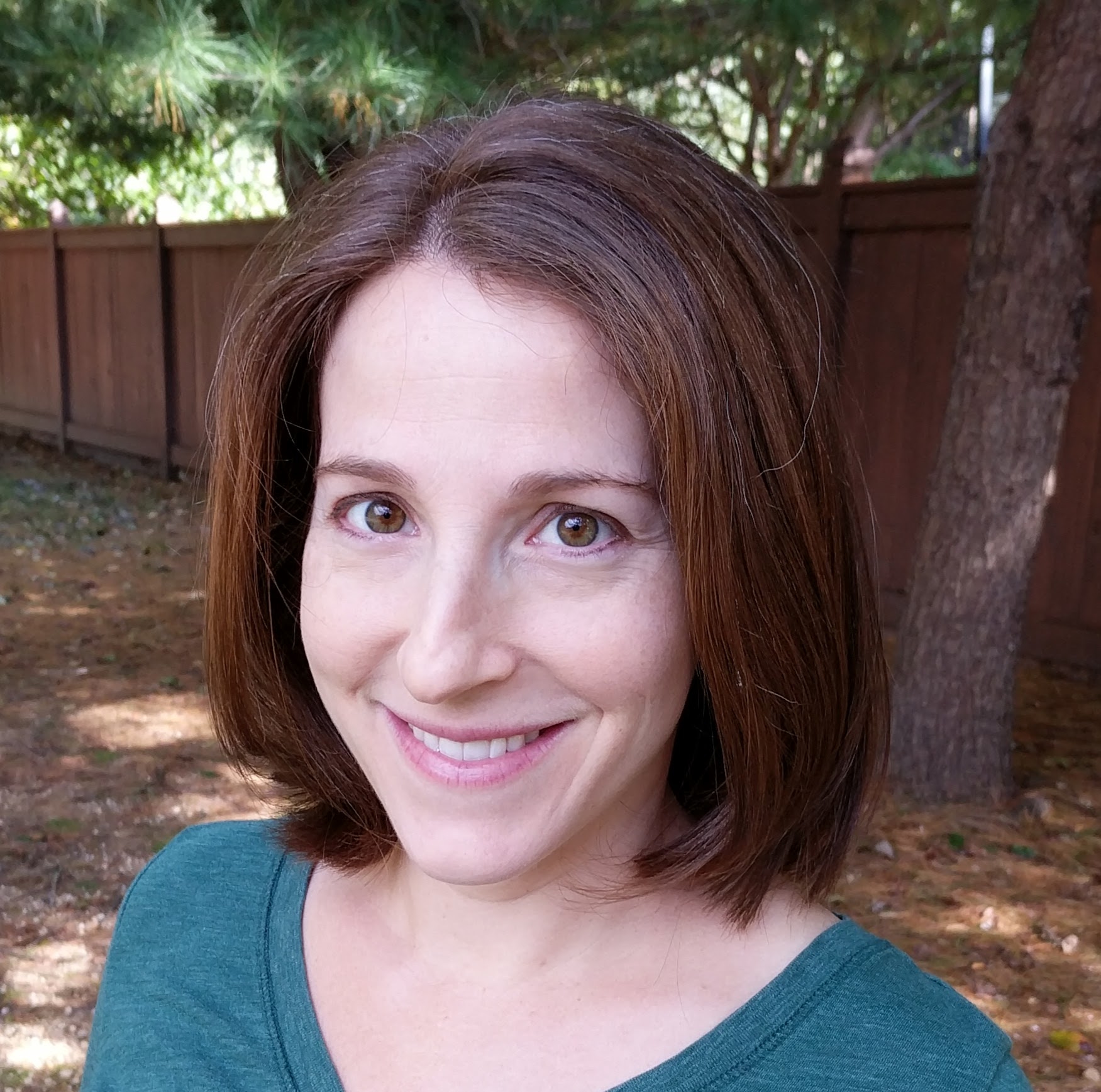 Woman with shoulder-length brown hair smiling outdoors, wearing a teal top, with trees and a wooden fence in background.