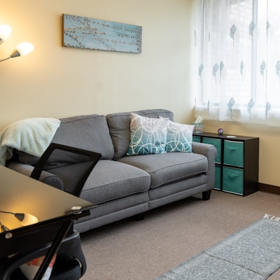 Living room with gray sofa, patterned pillow, side table, window with sheer curtains, and wall art.