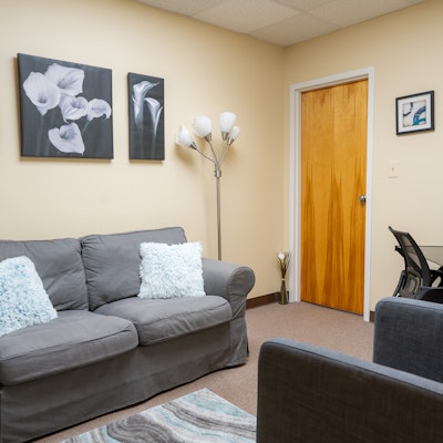 Living room with gray sofa, white pillows, wall art, a floor lamp, and a wooden door, beige walls, and carpeted floor.
