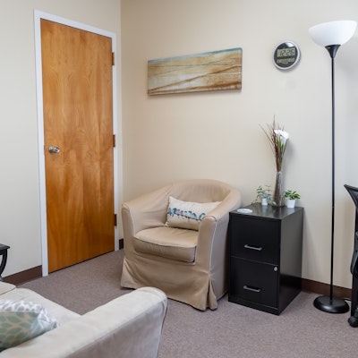 Corner of a room with a beige armchair, black side table, tall floor lamp, and wall decorations, including a painting and clock.