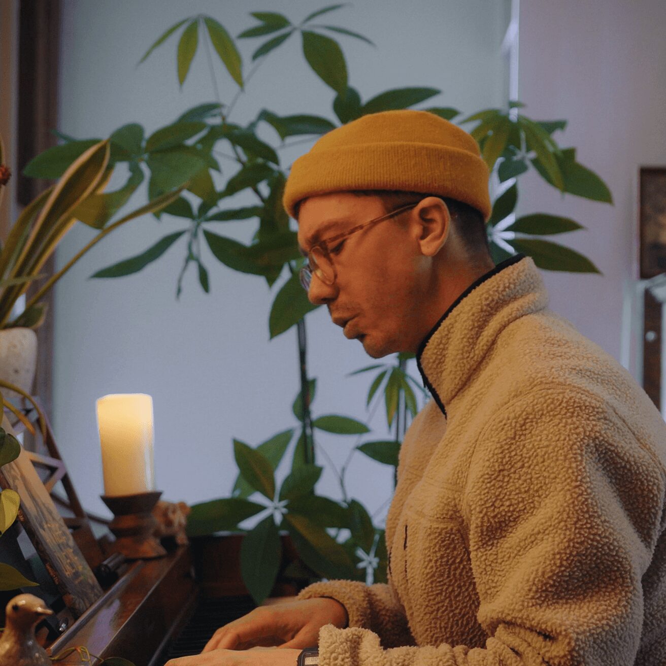 Person wearing glasses, yellow beanie, and beige fleece jacket, sitting indoors near a plant and candle.