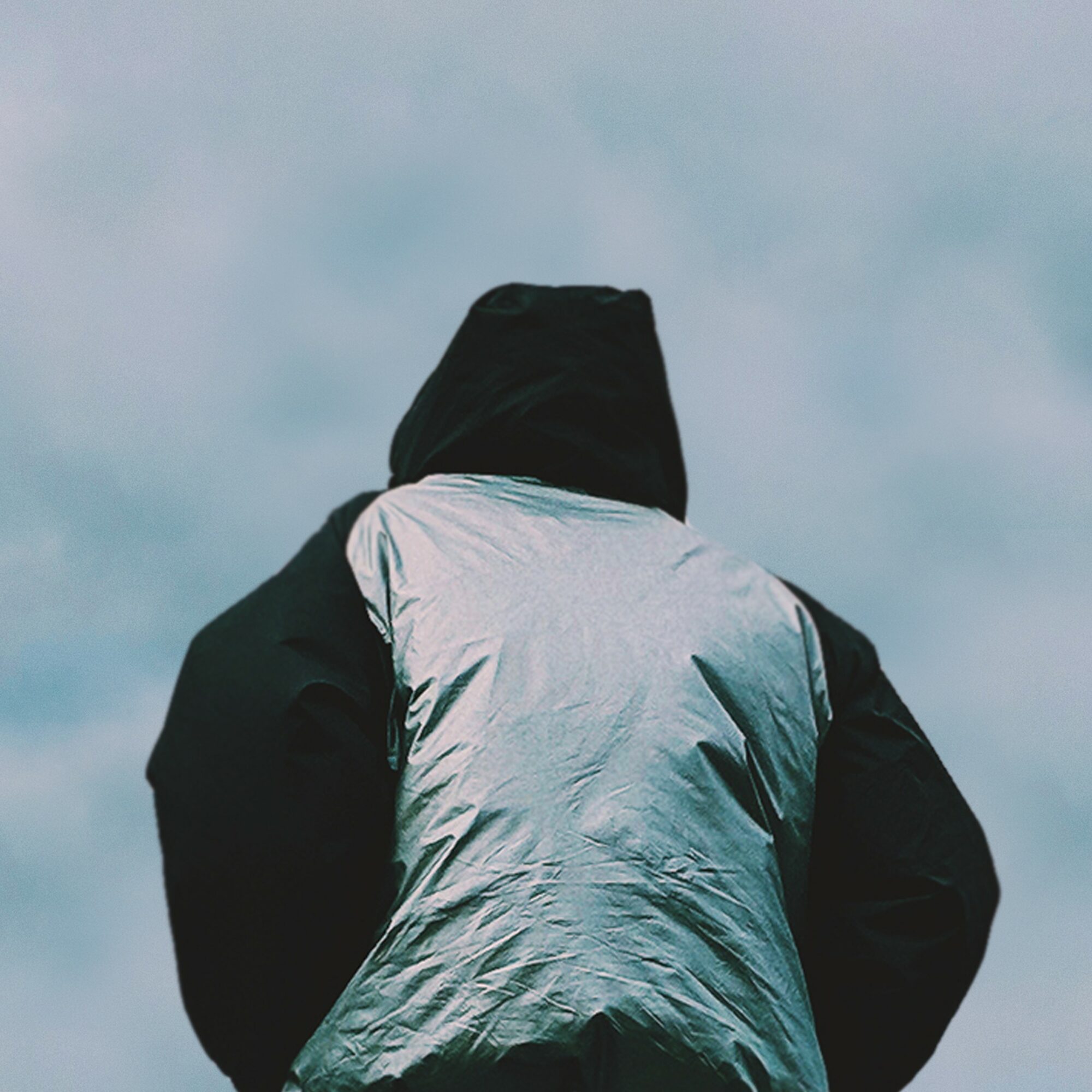 Person wearing a hooded jacket, facing away, with a cloudy sky background.