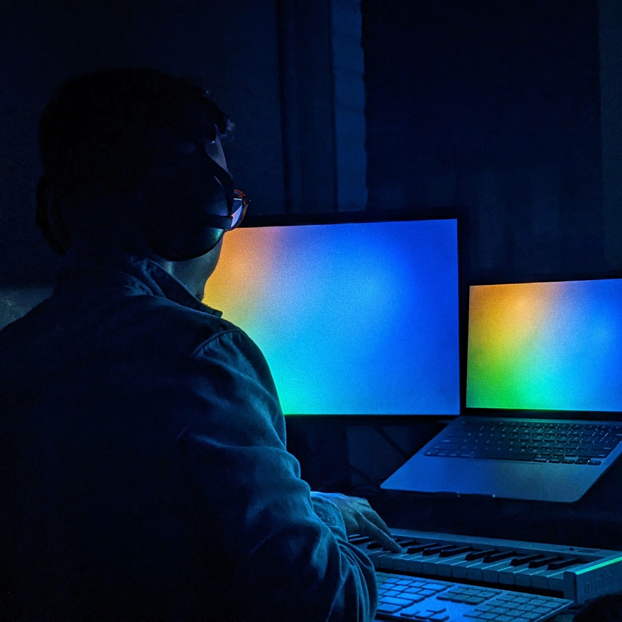 Person wearing glasses working at a desk with two monitors displaying colorful screens, in a dark room.