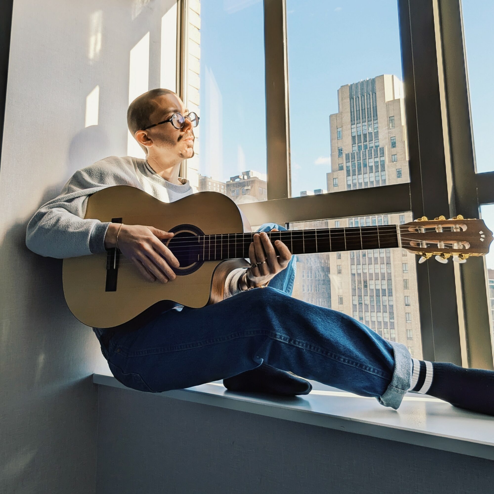 Person sitting on windowsill playing guitar, cityscape view outside window, sunlight streaming in, relaxed atmosphere.