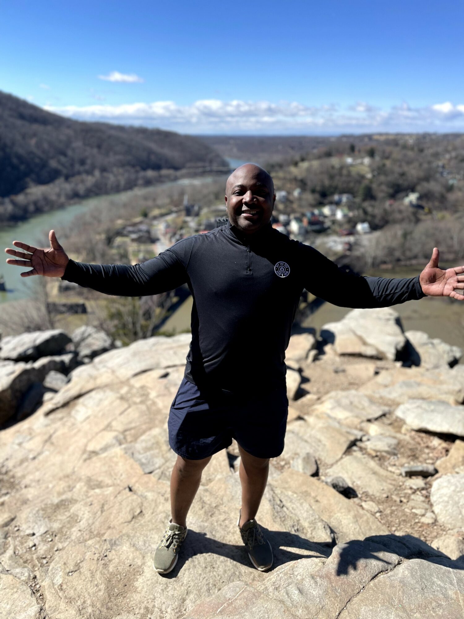 Man standing on rocks with arms outstretched, landscape of hills and sky in background.
