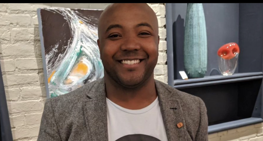 Smiling man in a gray blazer and white shirt standing indoors with artwork and vases in the background.