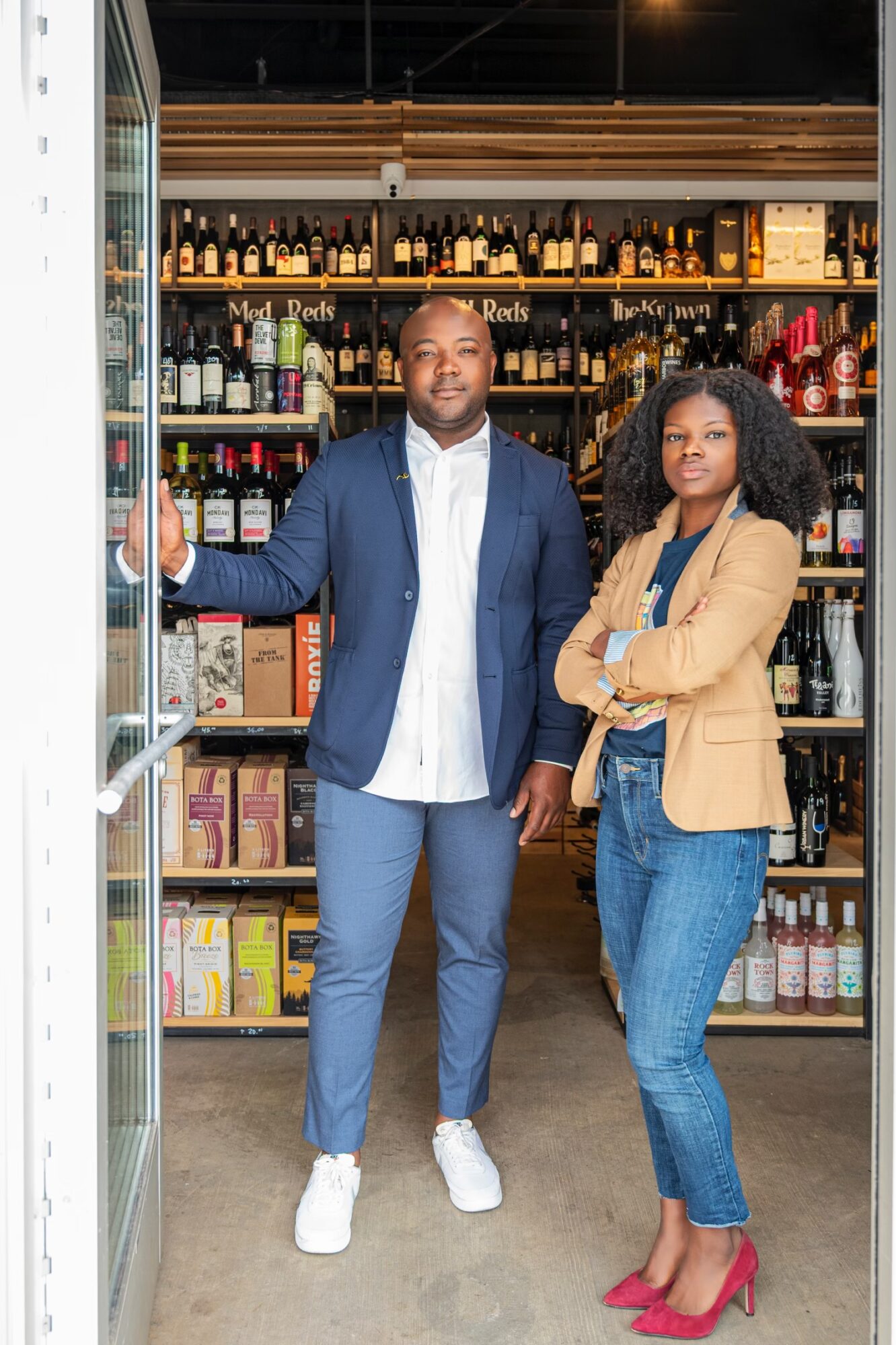 Two people stand in front of a store shelf filled with bottles and boxes, smiling at the camera.