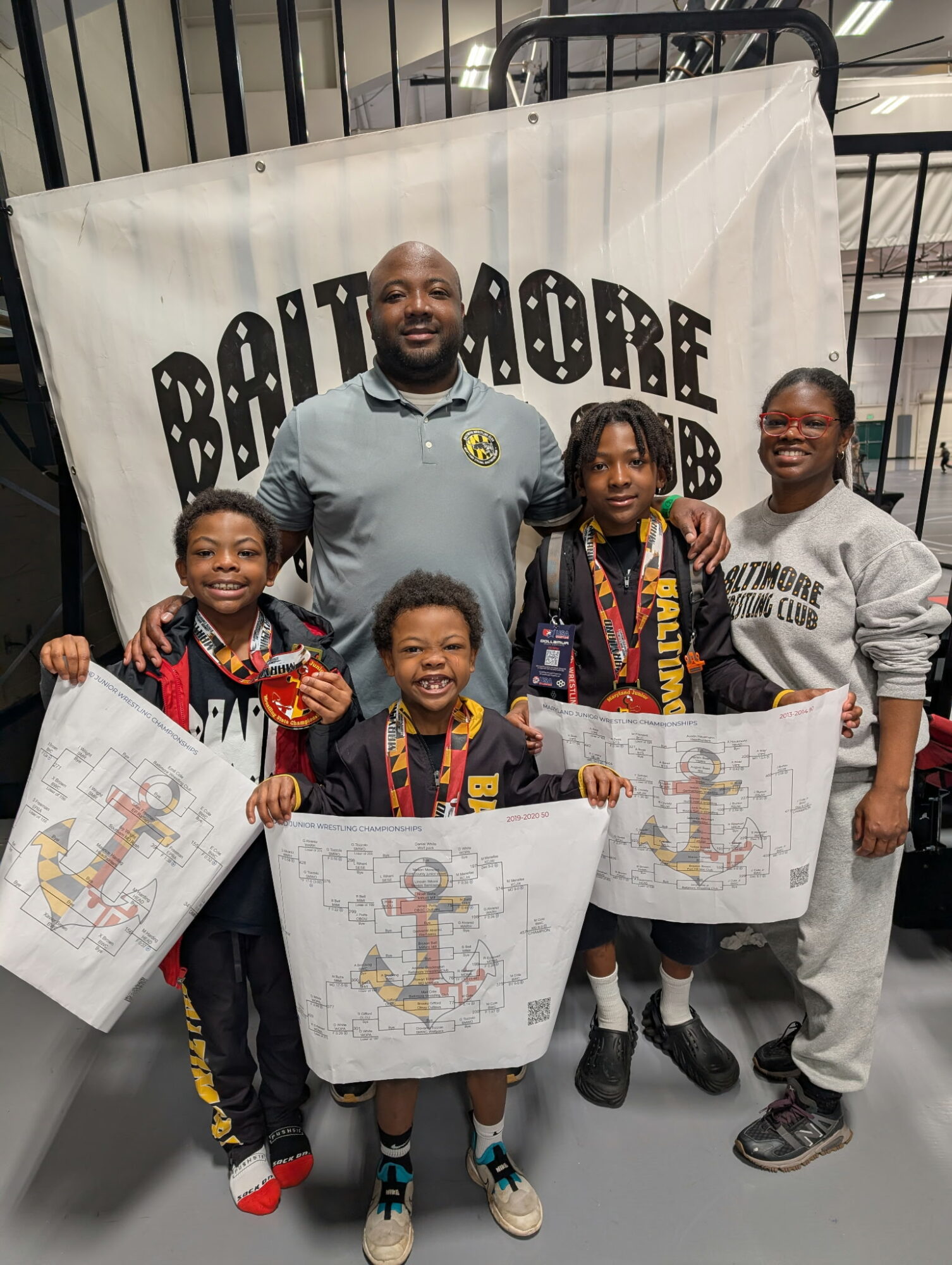 Group of five people, including children and an adult, standing in front of a Baltimore banner, holding large maps or posters.