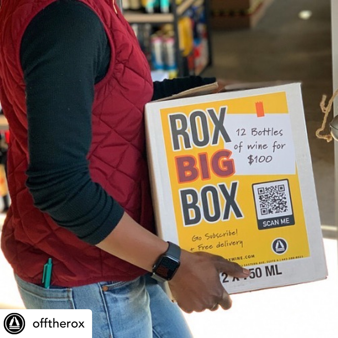 Person holding a box with a sign advertising bottles for $100, QR code, and subscription offer, in a store.