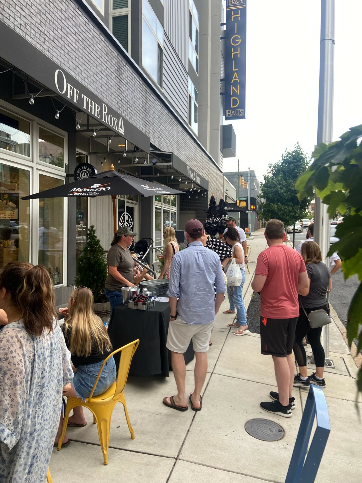 People walking on a sidewalk outside a building with a sign, some sitting on yellow chairs, and trees in the background.