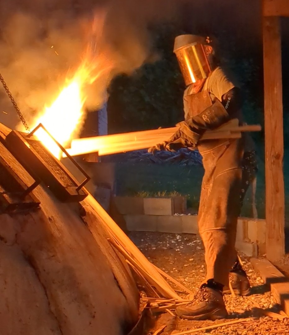 Person wearing protective gear working near a large fire with bright flames and smoke at night.