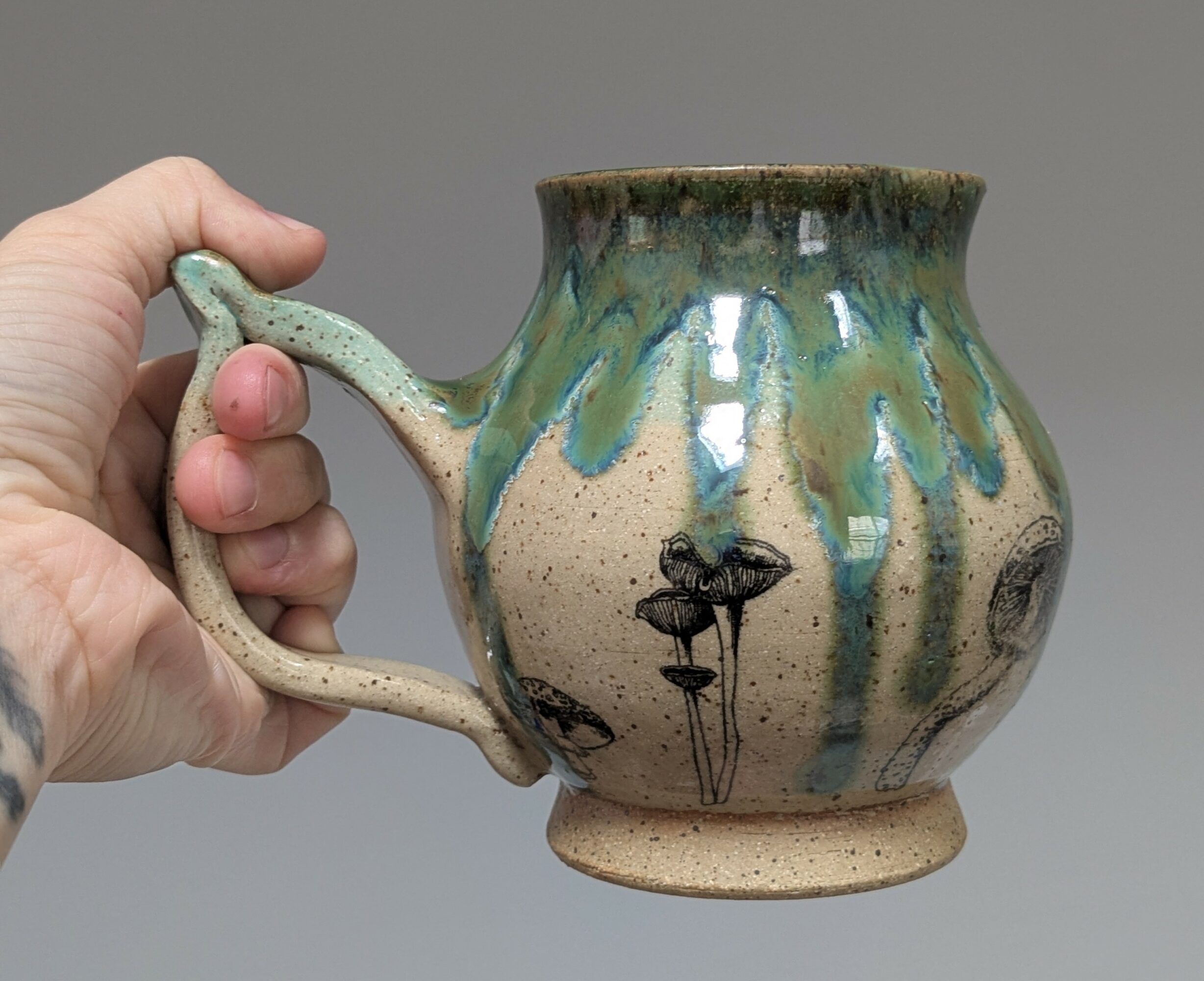 Hand holding a ceramic mug with a rounded body, decorated with green and blue glaze drips and black floral designs, against a gray background.