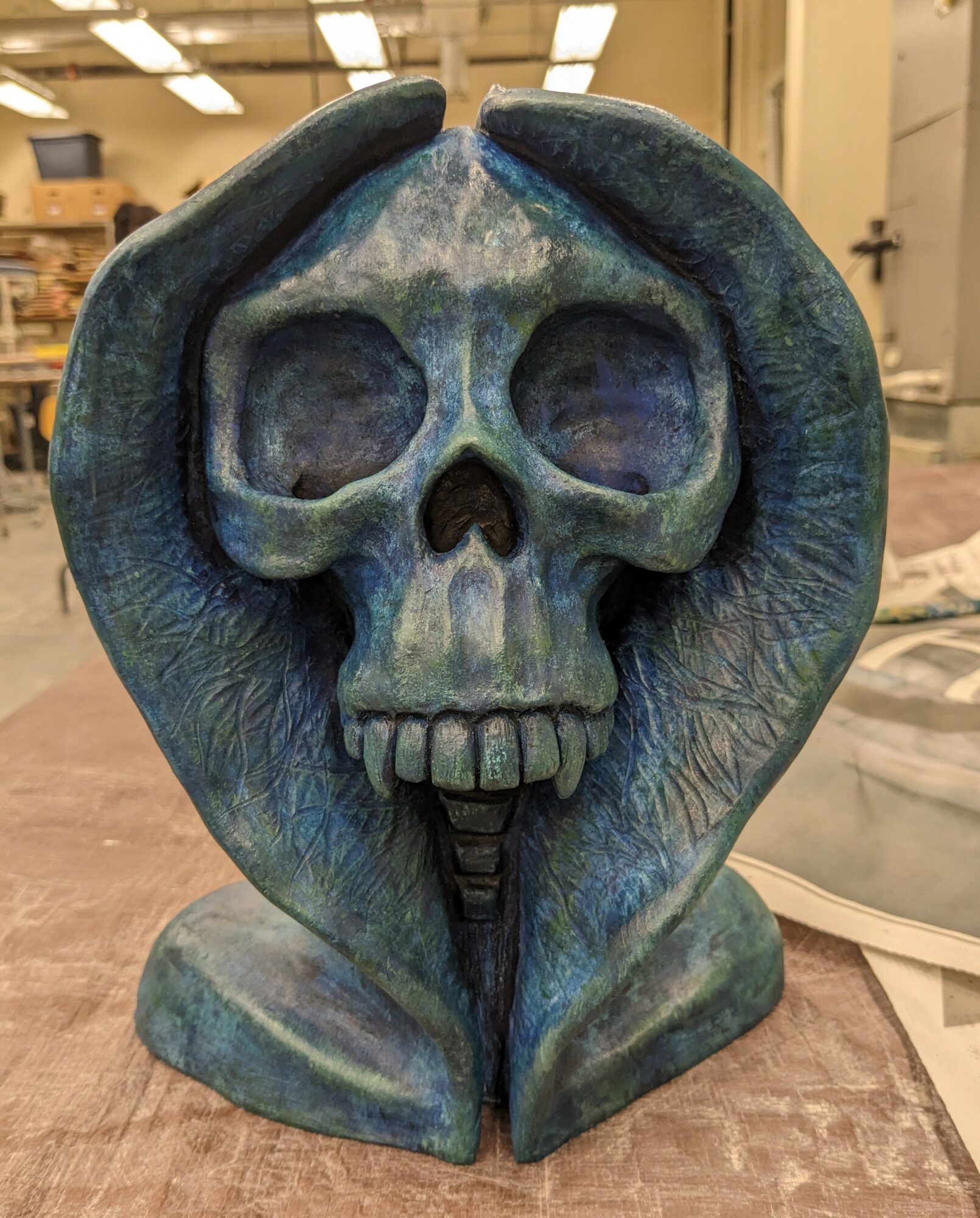 Blue skull sculpture with hands around its head, displayed indoors on a table, with a workshop background.
