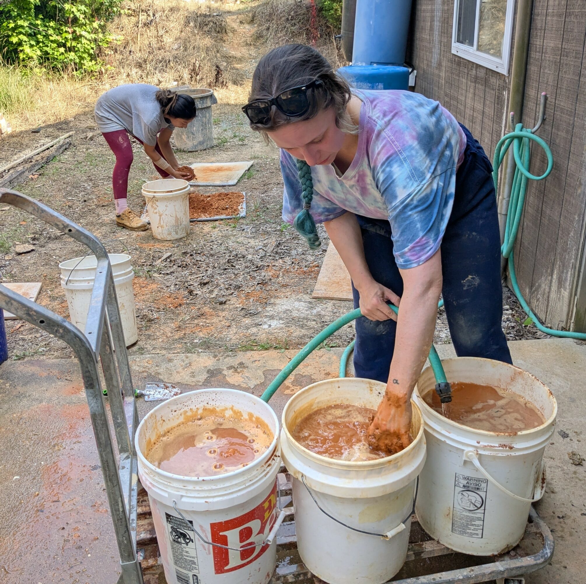 Two women working outdoors with buckets of muddy water, one bending over and the other using a hose, near a building.