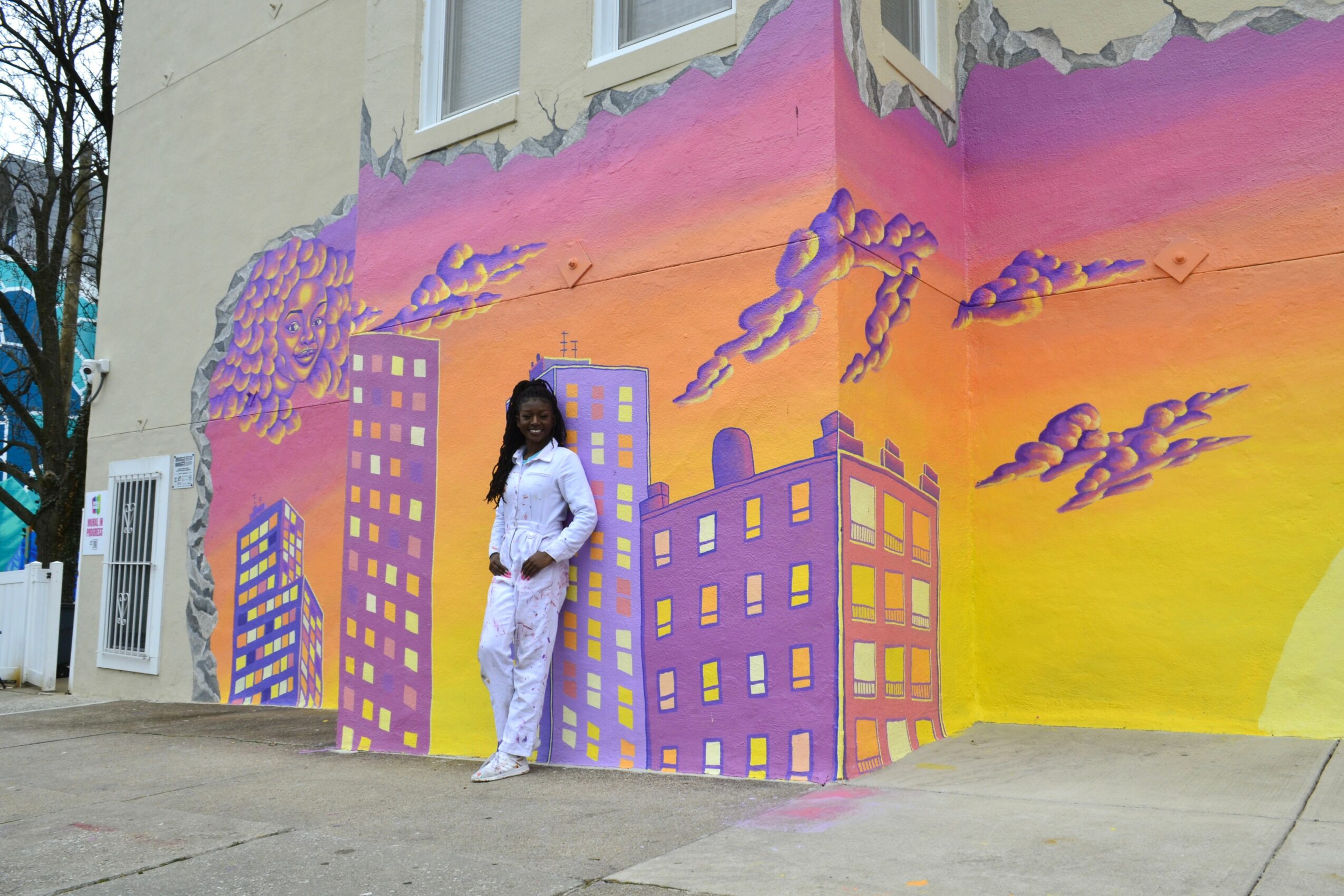 Person standing in front of colorful cityscape mural with buildings and clouds, sunset sky background.