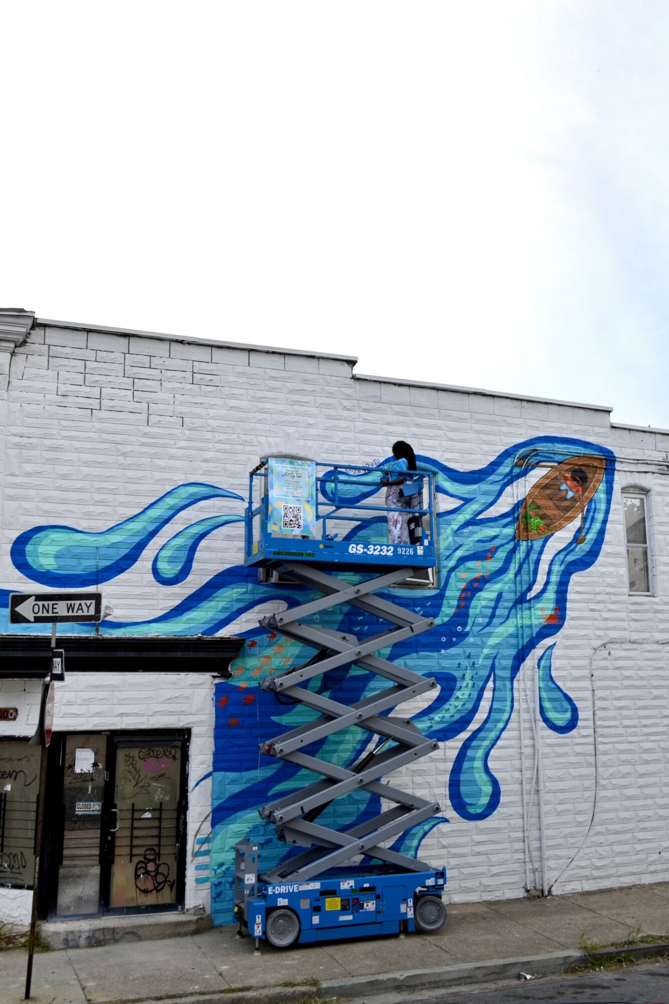 Person on a lift painting a mural of blue flowing hair with a fish on a white brick wall.