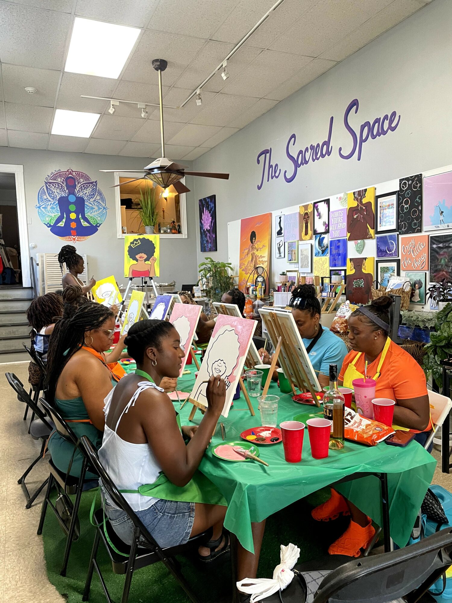 Group of children and adults painting at a table in an art studio with colorful artwork on the wall, and a sign reading 'The Sacred Space'.