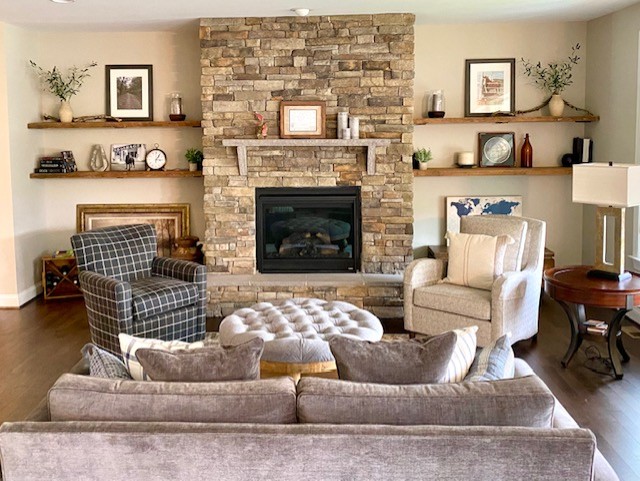 Living room with a stone fireplace, two shelves with decor, two armchairs, a sofa, and a round ottoman.