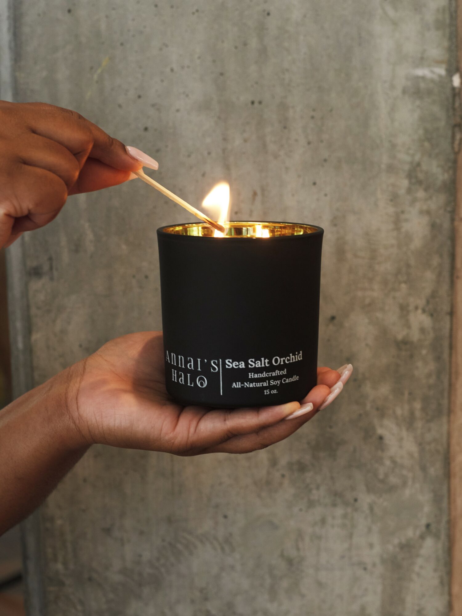 Person holding a black candle labeled 'Sea Salt Orchid' with a lit match near the flame, against a concrete background.