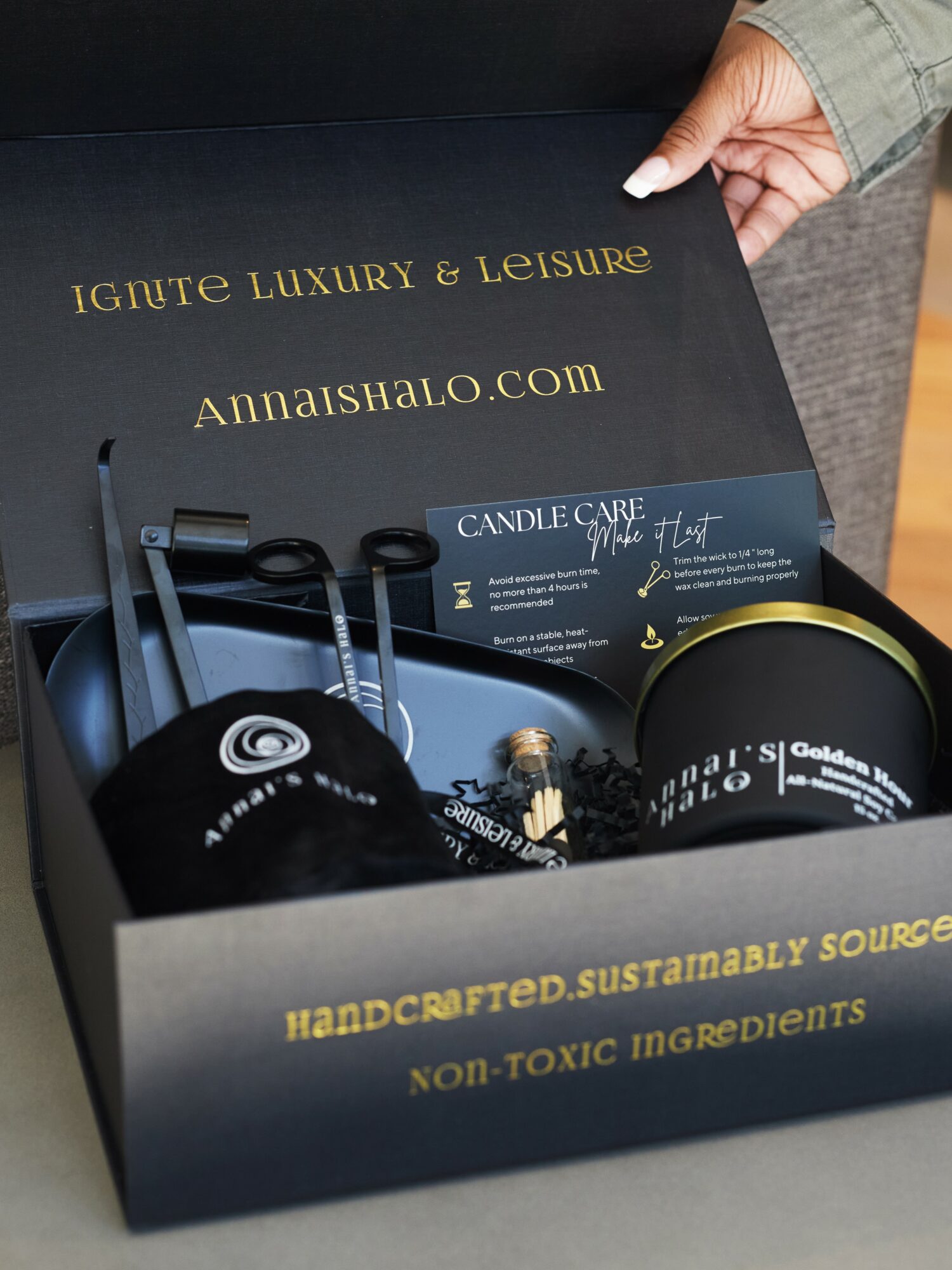 Open black box with gold text, containing candles and candle care card, held by a person in a green jacket.