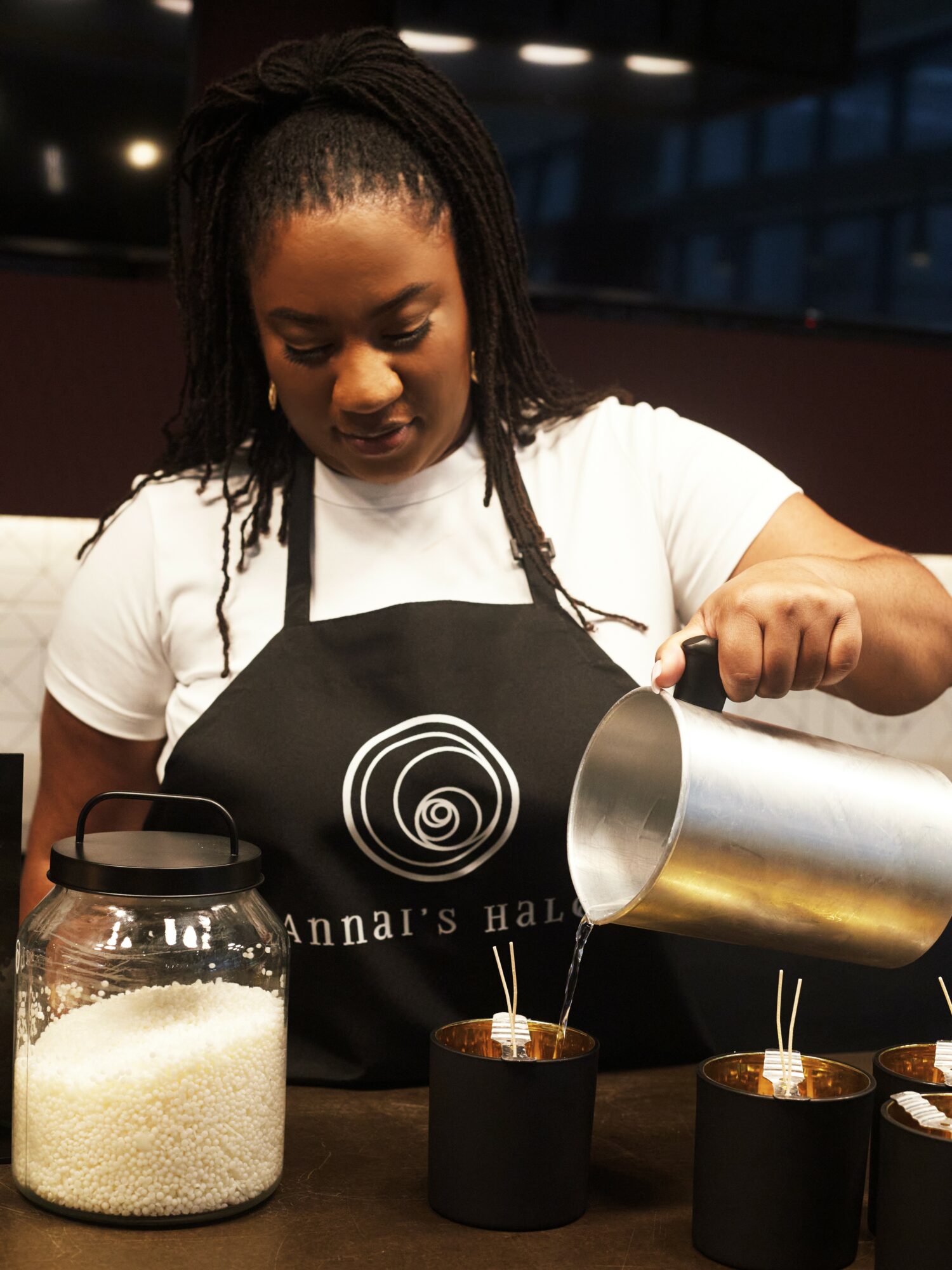 Woman pouring liquid into cups, wearing an apron, in a kitchen or cafe setting.