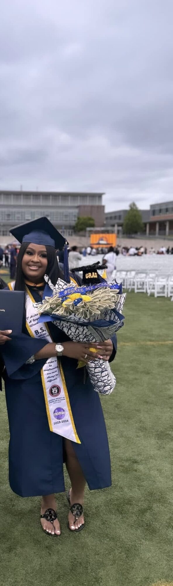 Person in graduation gown holding a bouquet of flowers outdoors on a cloudy day.