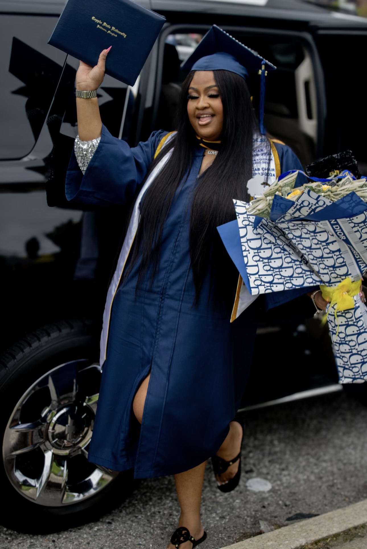 Graduate in cap and gown holding a diploma and flowers, smiling near a vehicle, celebrating graduation.