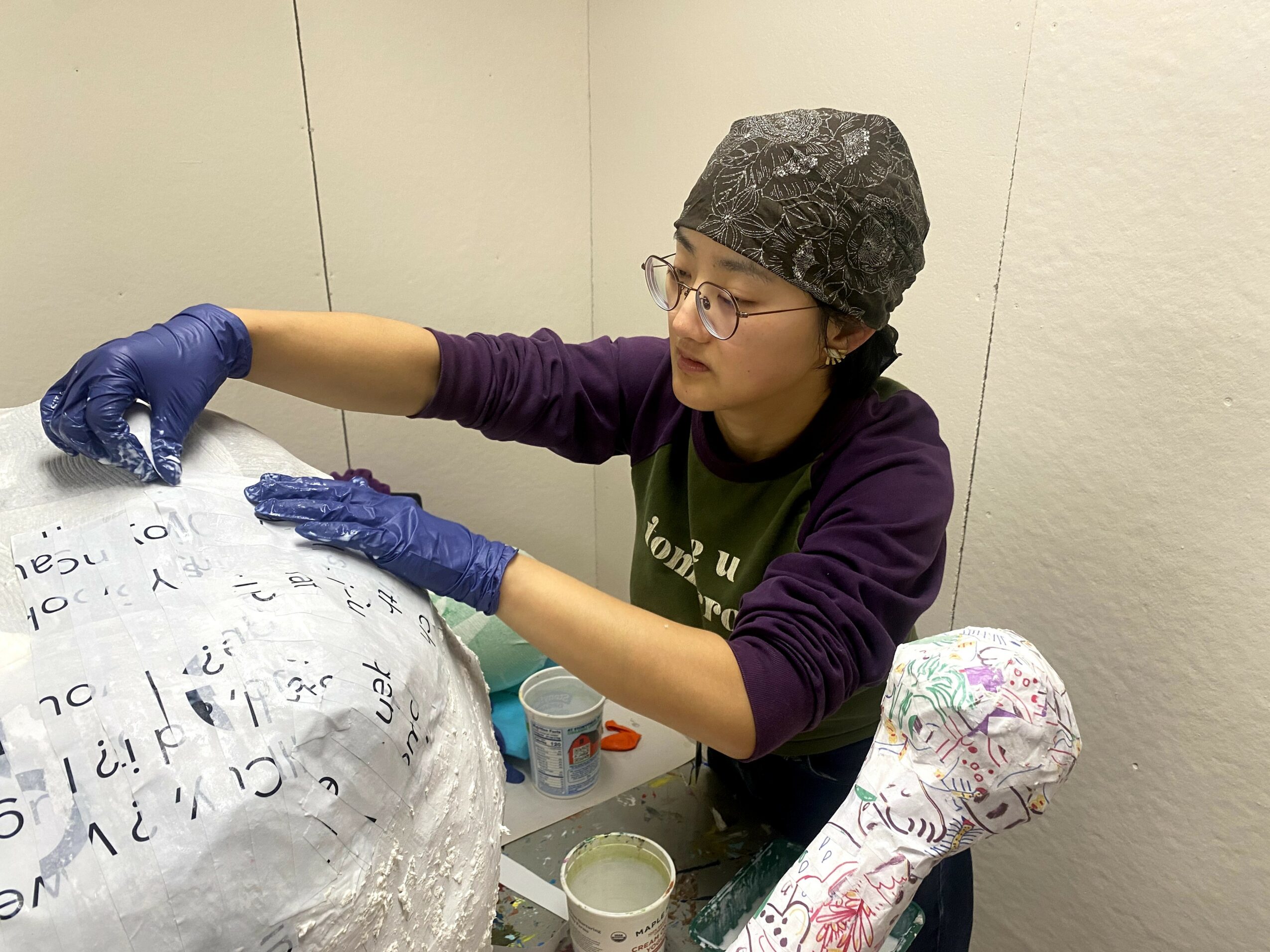 Person wearing glasses, a gray head covering, and purple gloves, working on a large object with writing on it, in a small room.