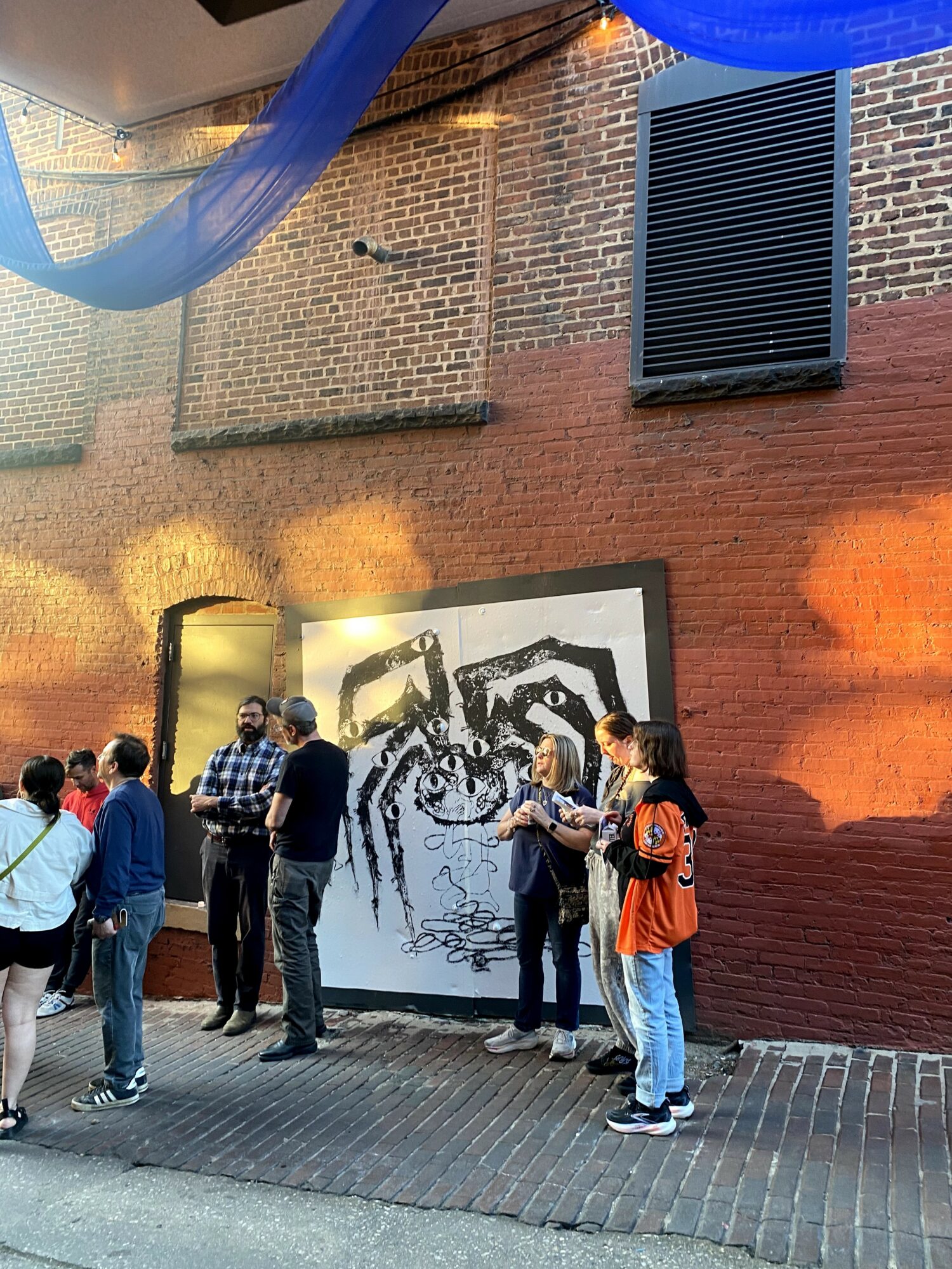 Group of people standing outside near a large black and white artwork on a brick wall, with a blue fabric decoration overhead.