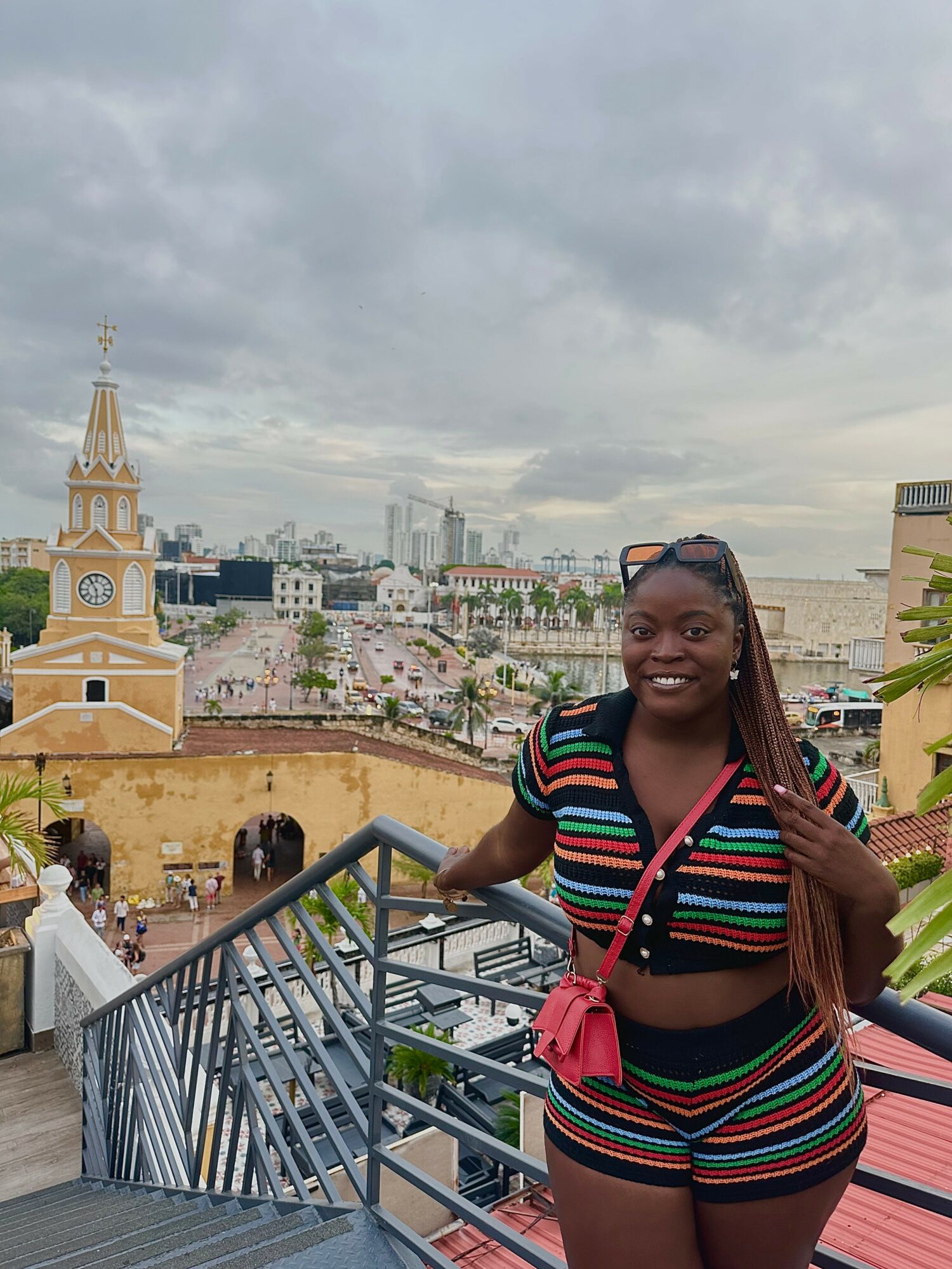 Smiling woman in colorful striped outfit standing on a balcony with cityscape and historic buildings behind her.