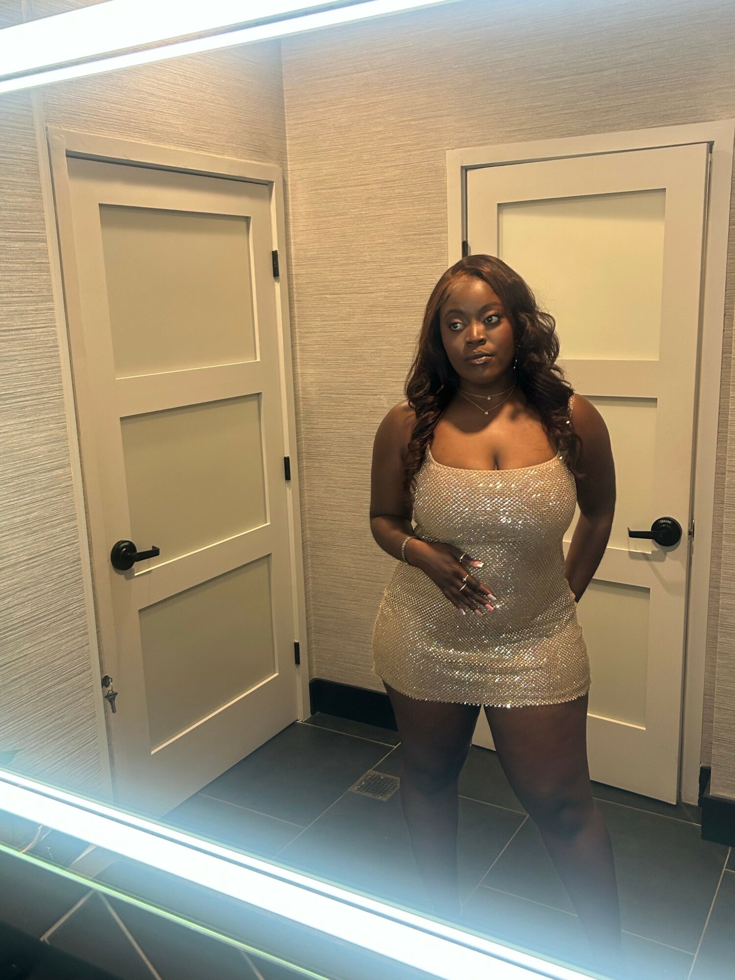 Woman in a sparkly dress standing in front of two closed doors, looking at the camera, in a room with beige walls.