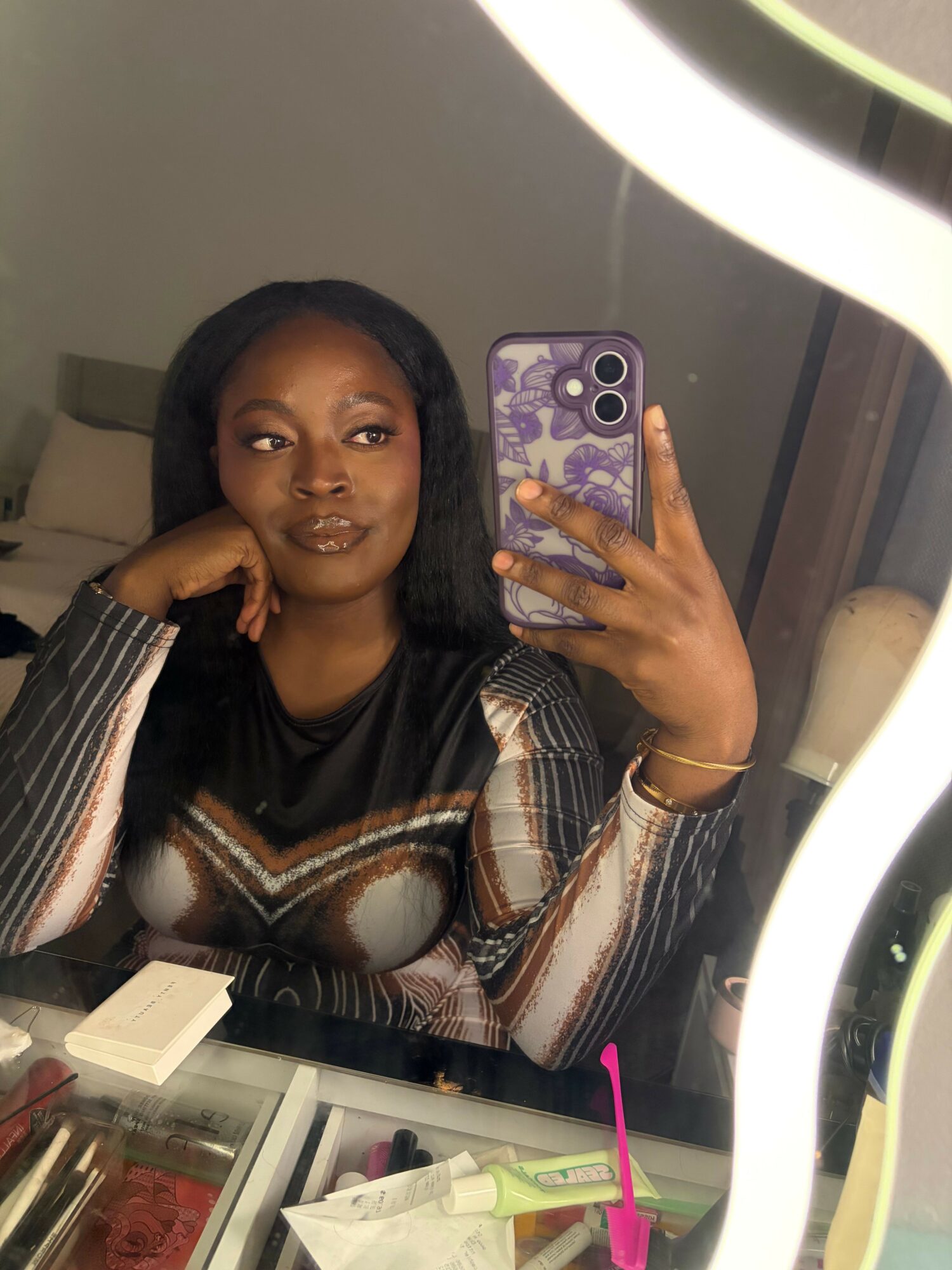 Woman taking a mirror selfie with a smartphone, sitting at a desk with various items, wearing a patterned top.
