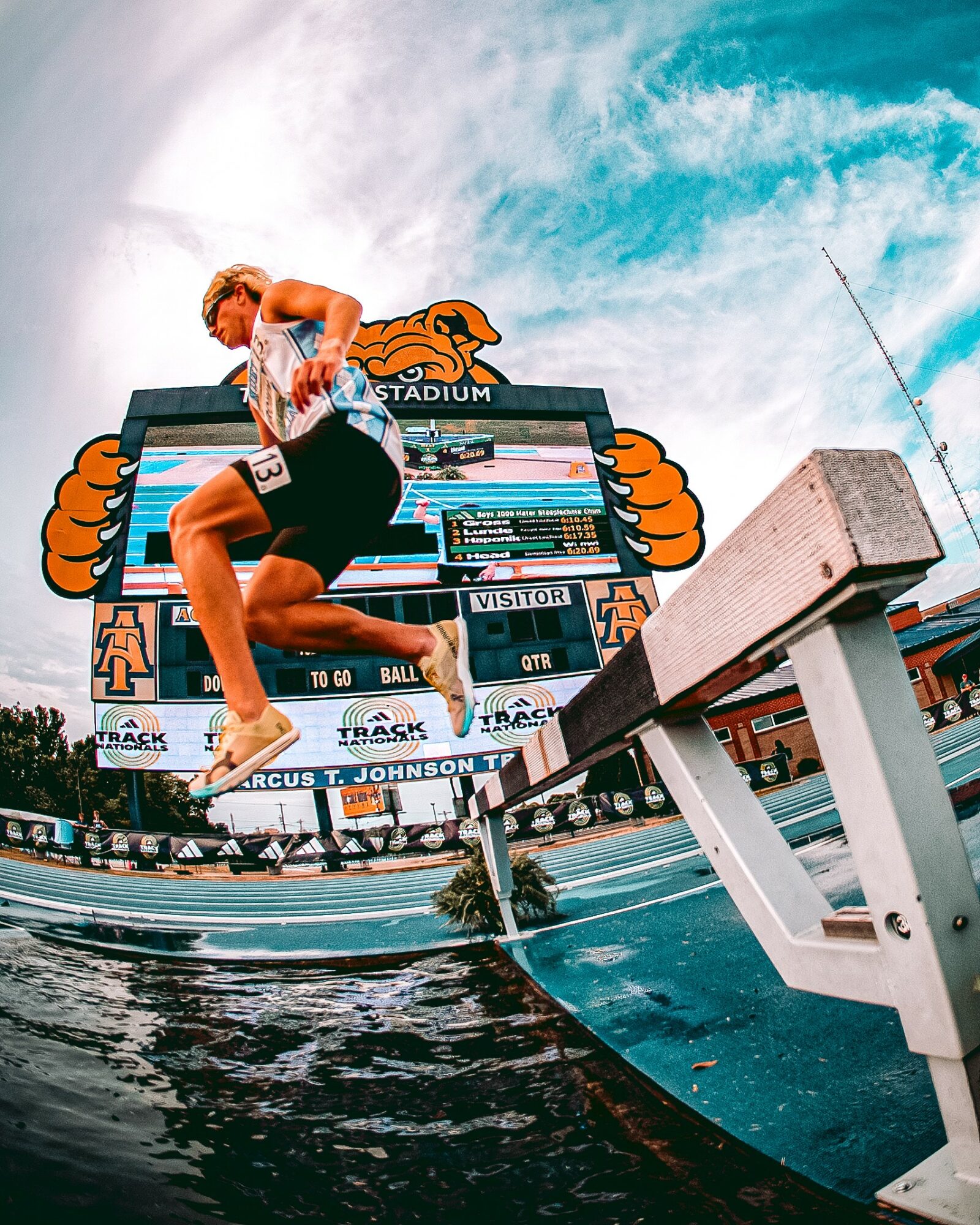 Person jumping off a dock into water at a stadium with a large screen and blue sky, captured from below.