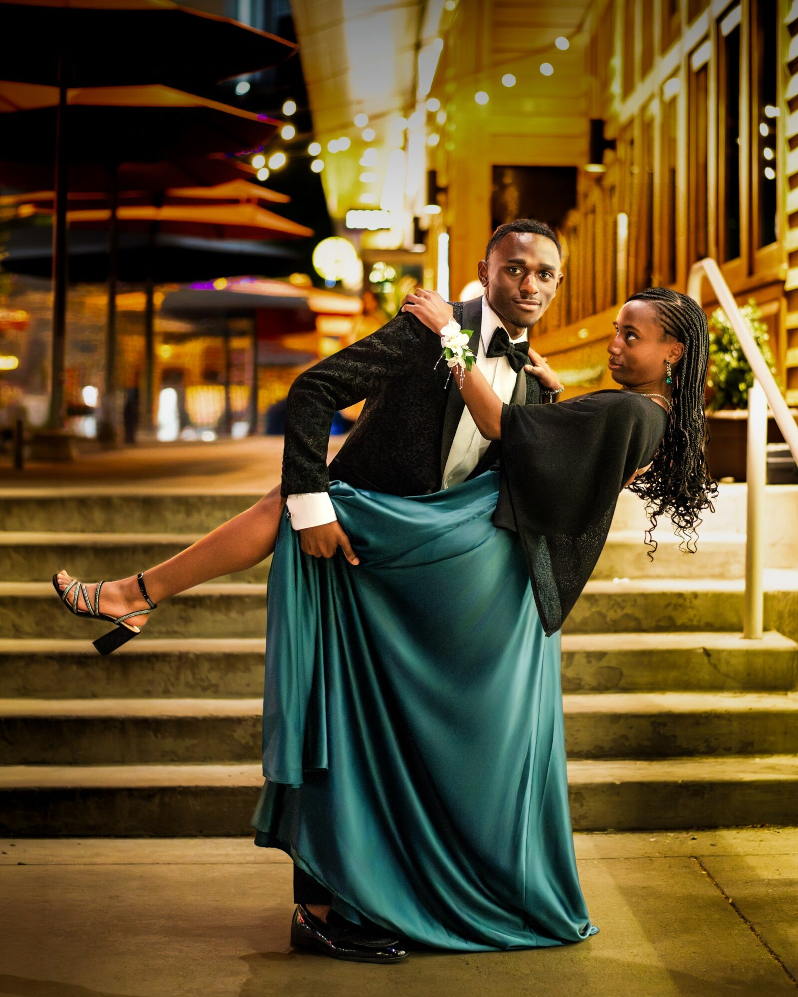 A couple dancing on stairs at night, the man in a tuxedo holding the woman in a long teal dress, illuminated by warm lights.