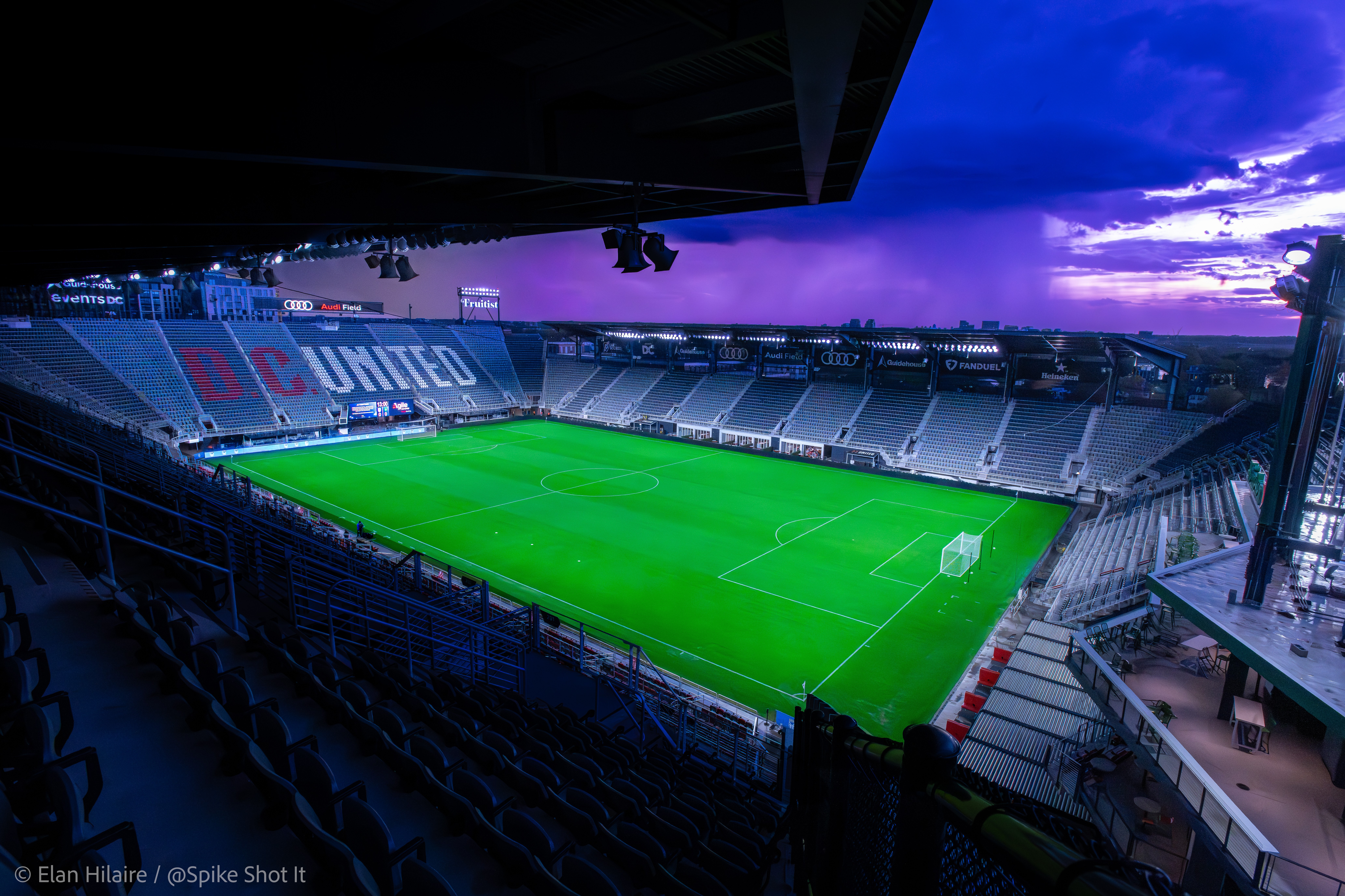 Empty soccer stadium with a green field, seating, and a sky with purple and blue clouds.