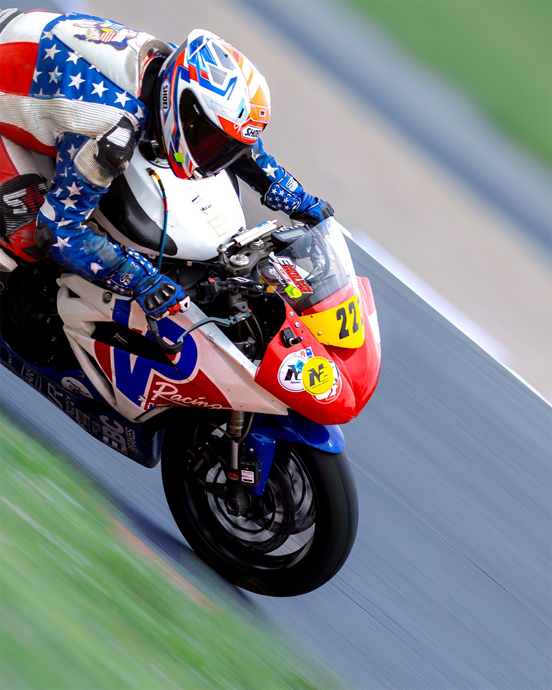 Motorcycle racer in blue, white, and red gear leaning into a turn on a race track, with a red and yellow number plate.