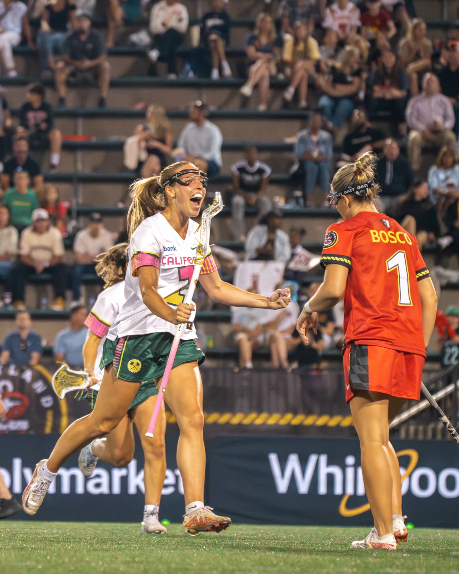 Two female soccer players celebrating on the field, one in a white jersey and the other in a red jersey, with spectators in the background.